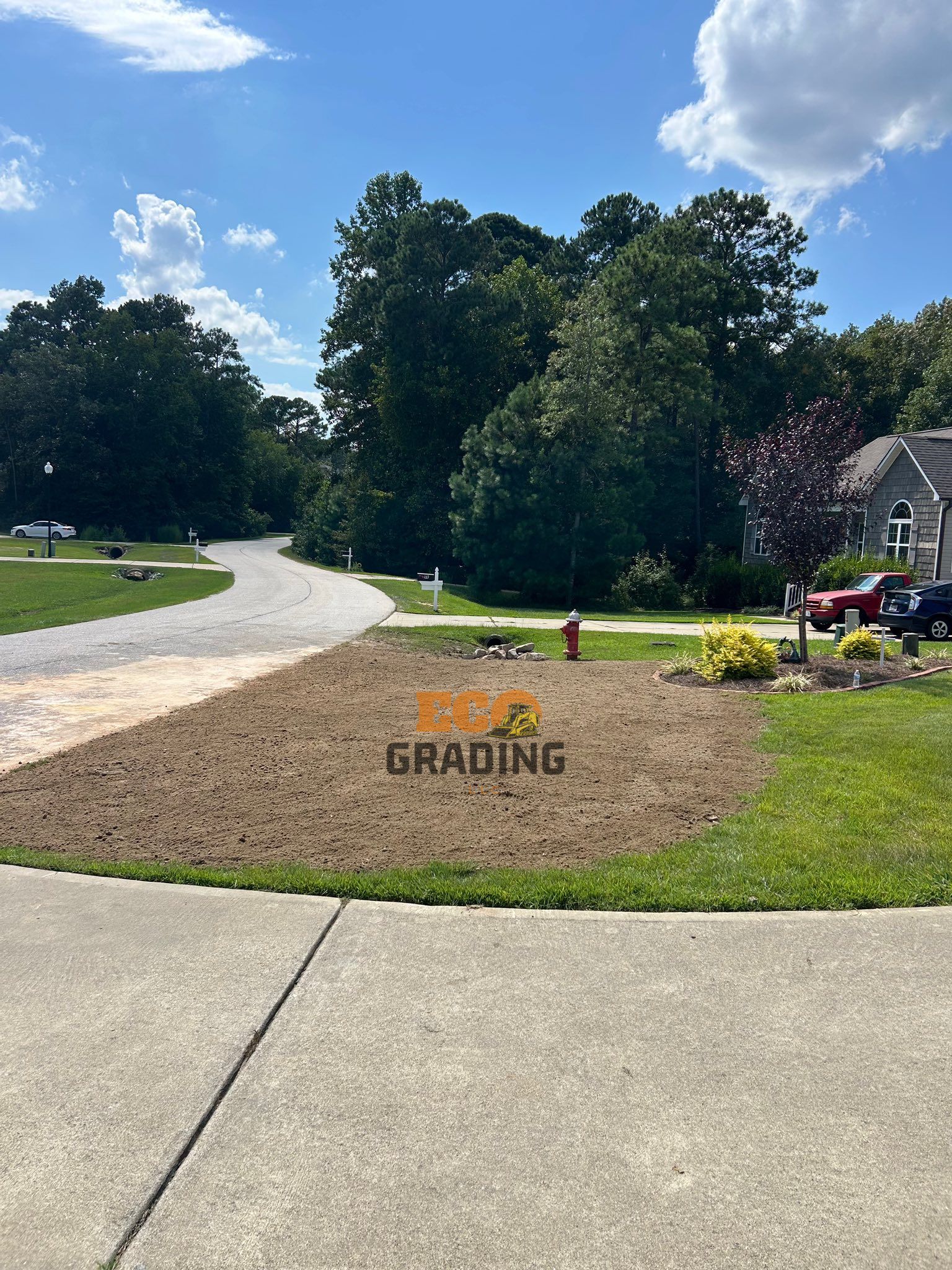 A graded yard with dirt, a driveway, grass, trees, and a house under a blue sky.