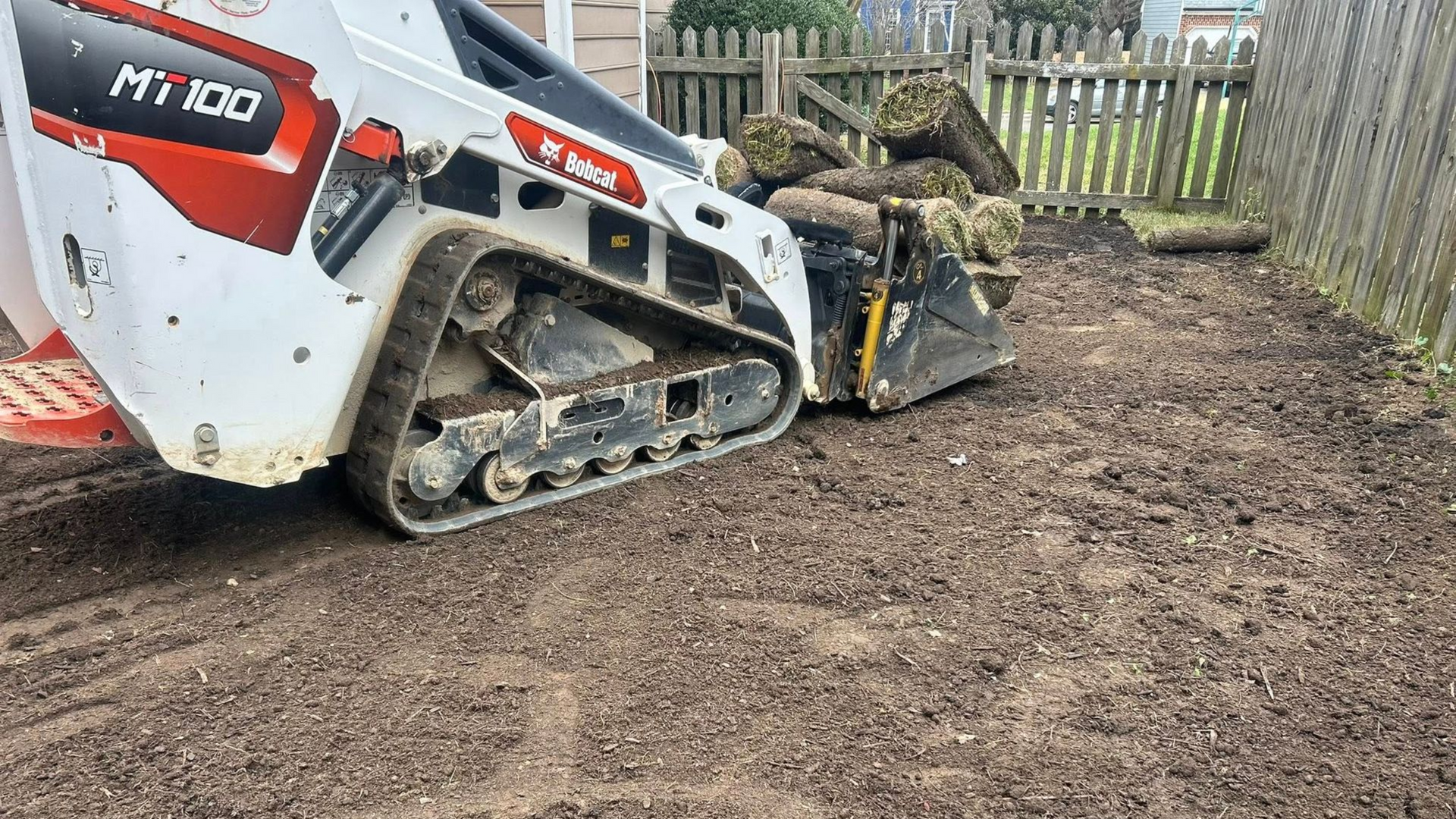 A Bobcat MT100 track loader is moving dirt in a yard next to a fence.