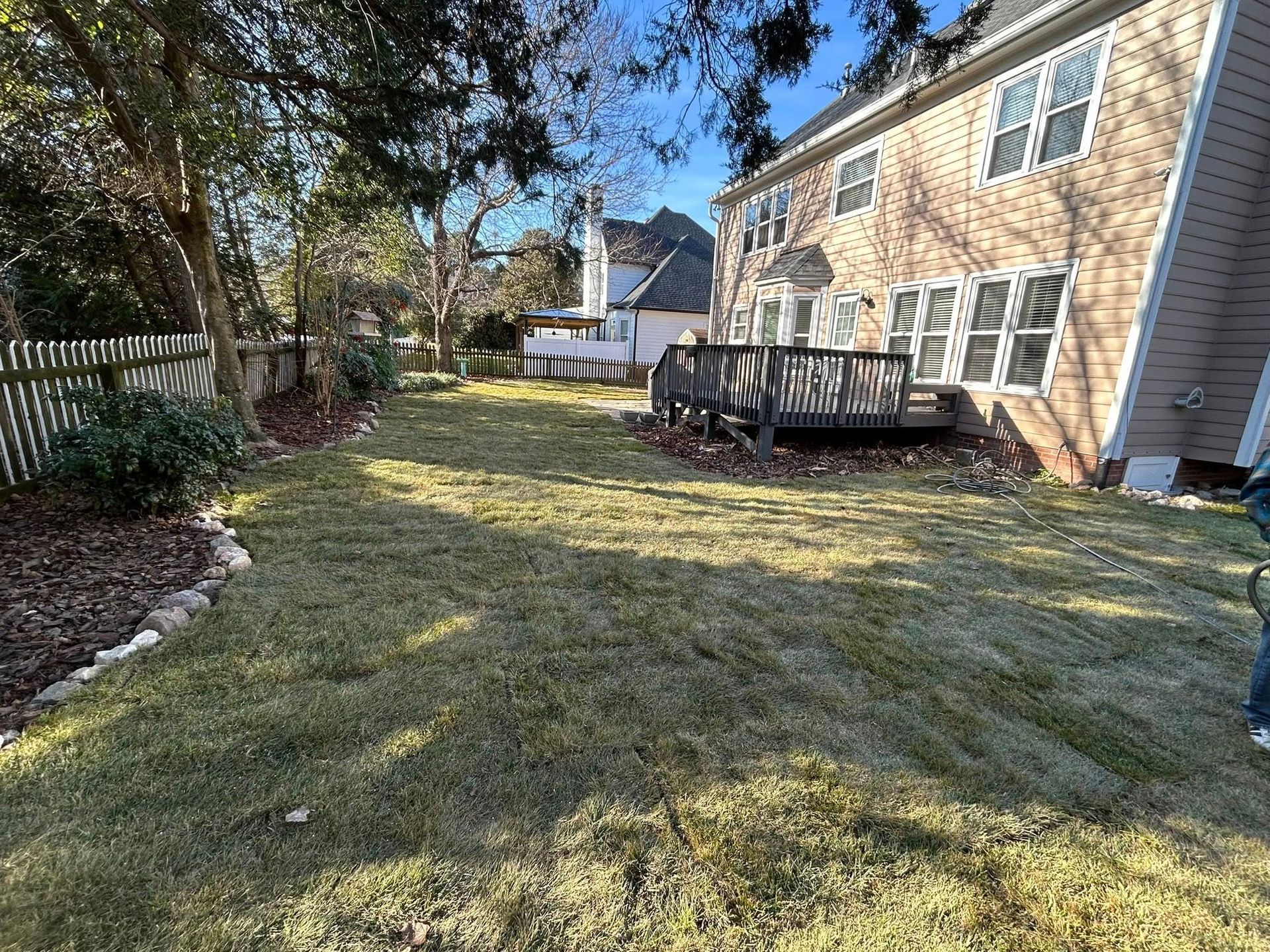 Backyard view of a house with a wooden deck, grass, and a fence. Sunny day, shadows on the grass.