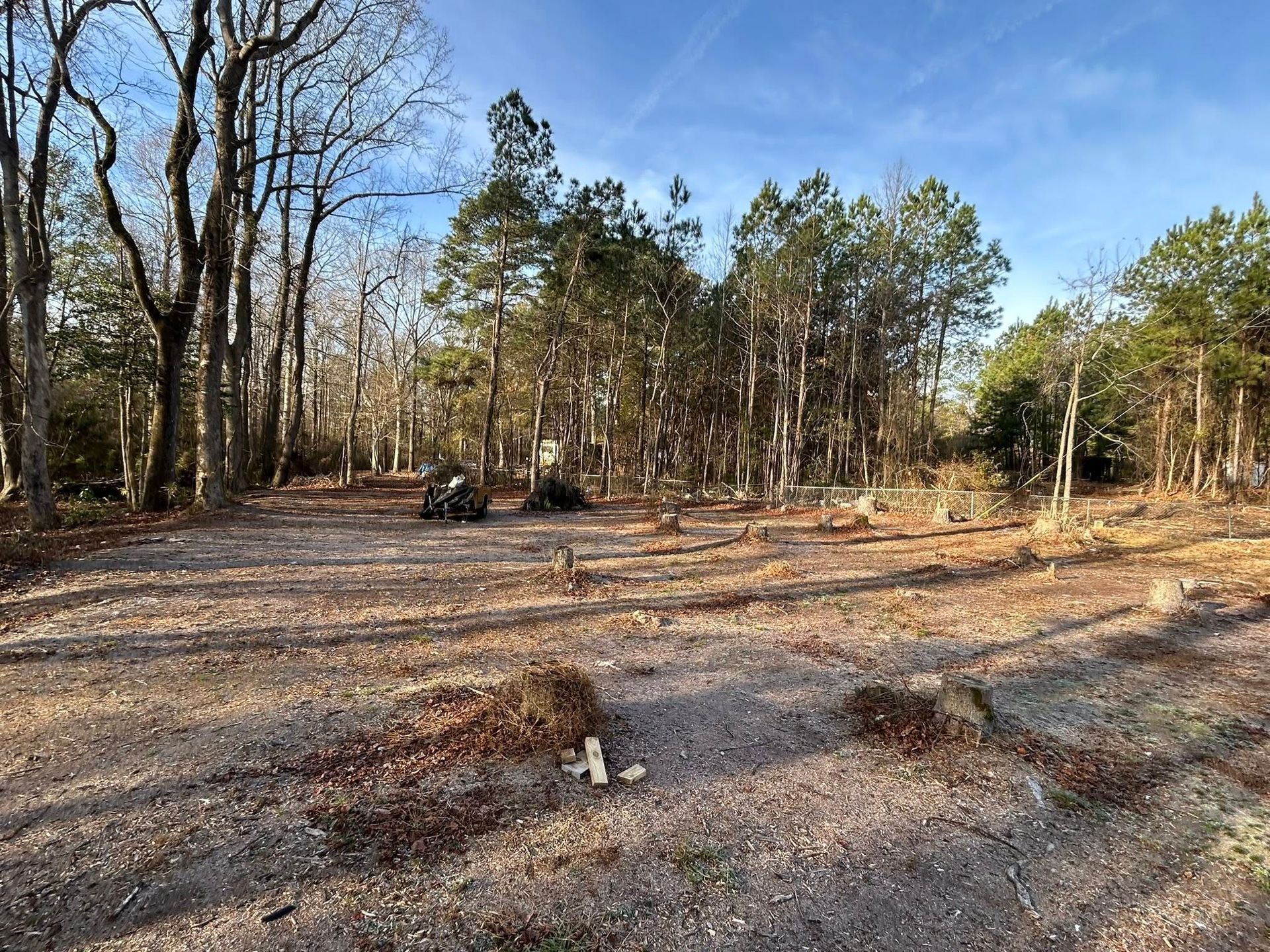Cleared dirt lot with scattered debris and trees in the background under a blue sky.