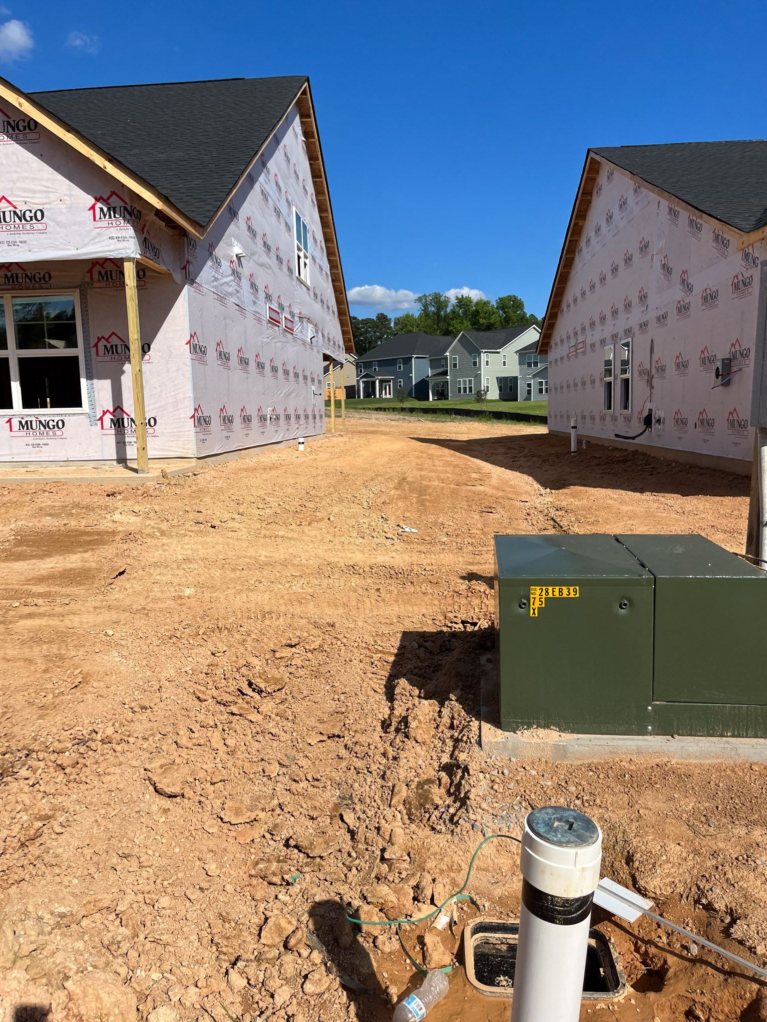 Two unfinished houses, separated by a dirt path, with a green electrical box and pipes in the foreground.