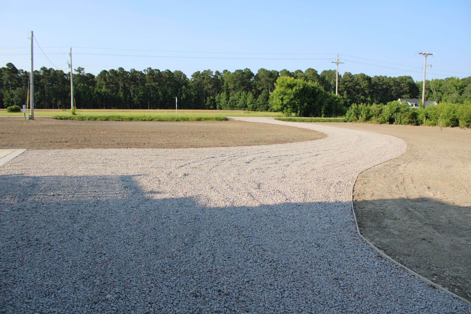 Gravel path curves through a graded area toward a tree line under a blue sky.