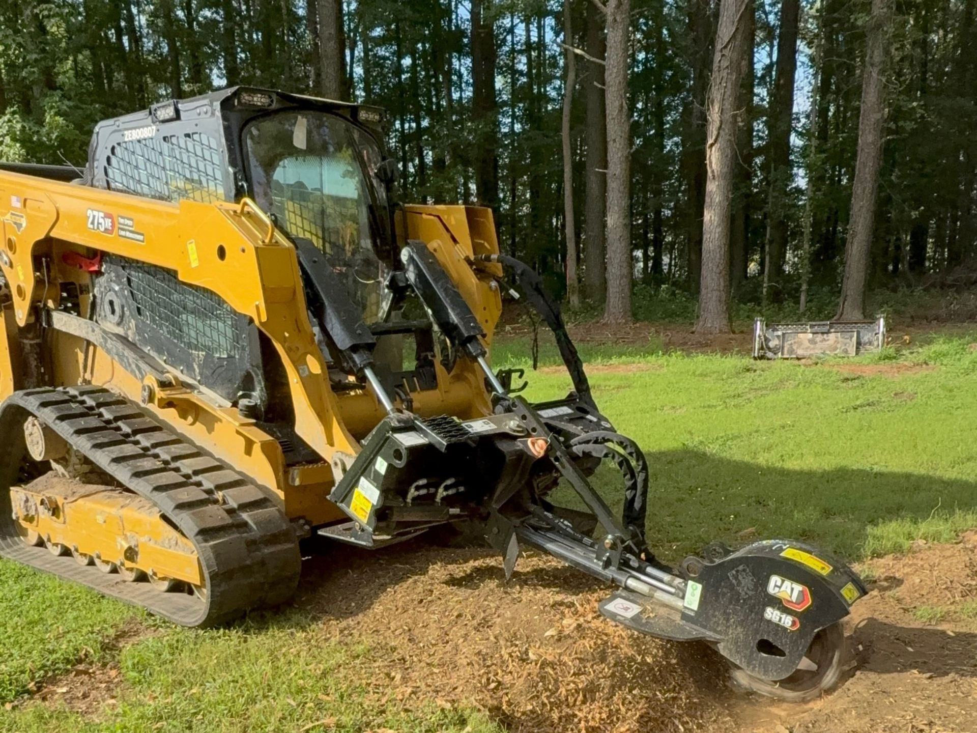 Yellow skid steer mulching grass and brush on a lawn, trees in background.