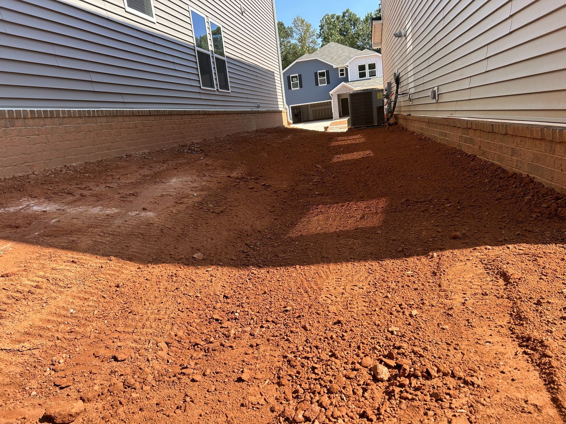 Dirt-covered ground between two houses with light-colored siding. Distant blue house visible.