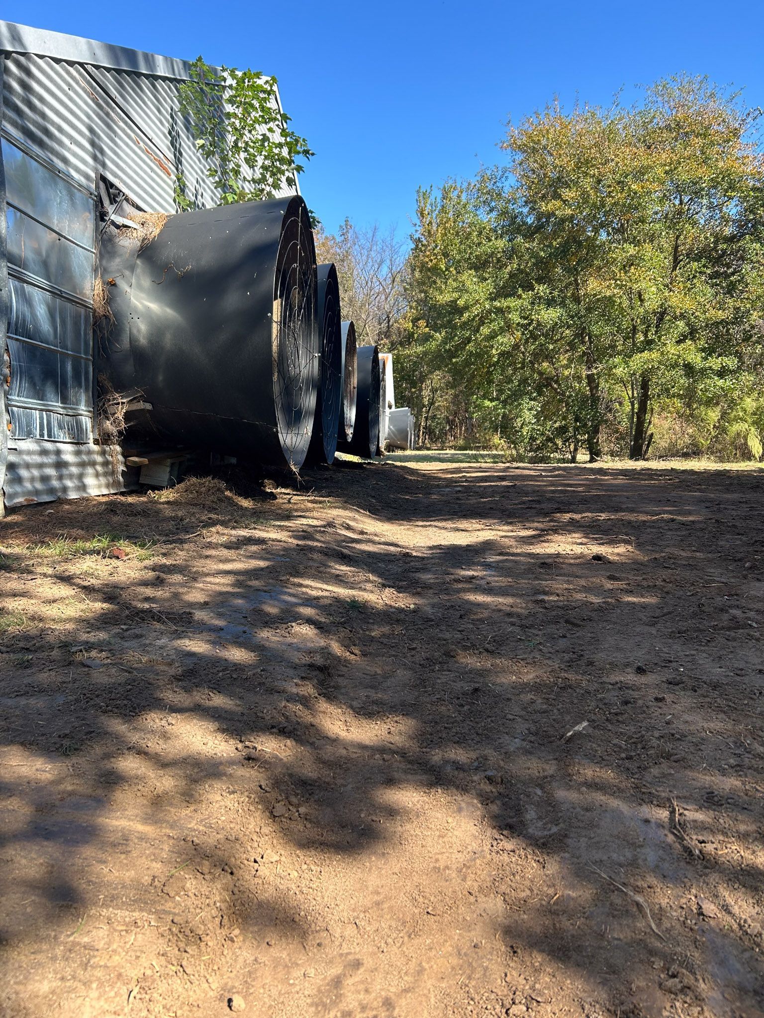 Dark containers sit against a building on a dirt lot, trees in background, sunny day.