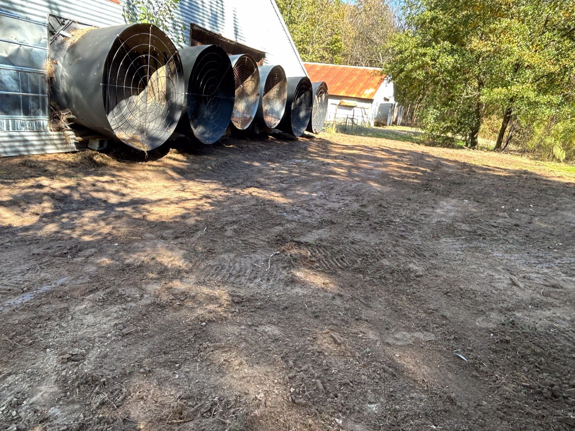 Five large, dark, cylindrical exhaust fans line the side of a white building, dirt ground in front, trees in the distance.
