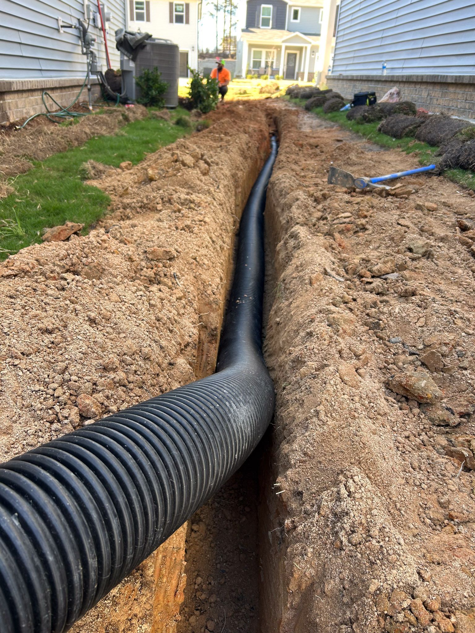 Black corrugated drainage pipe in a deep trench. Dirt and gravel surrounding. Person in the background.