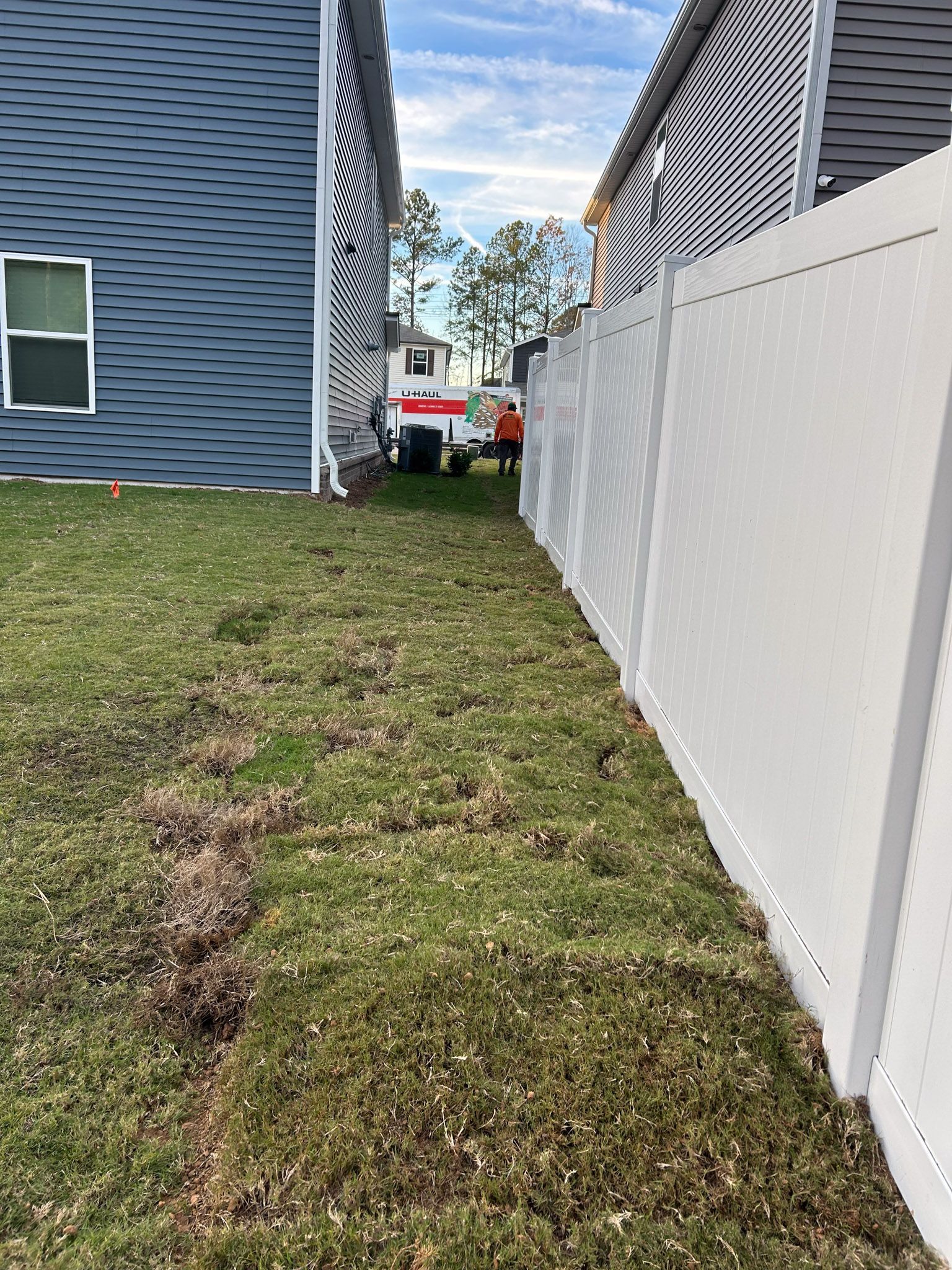 Backyard with white fence, two-story house with blue siding, and work truck in the distance.