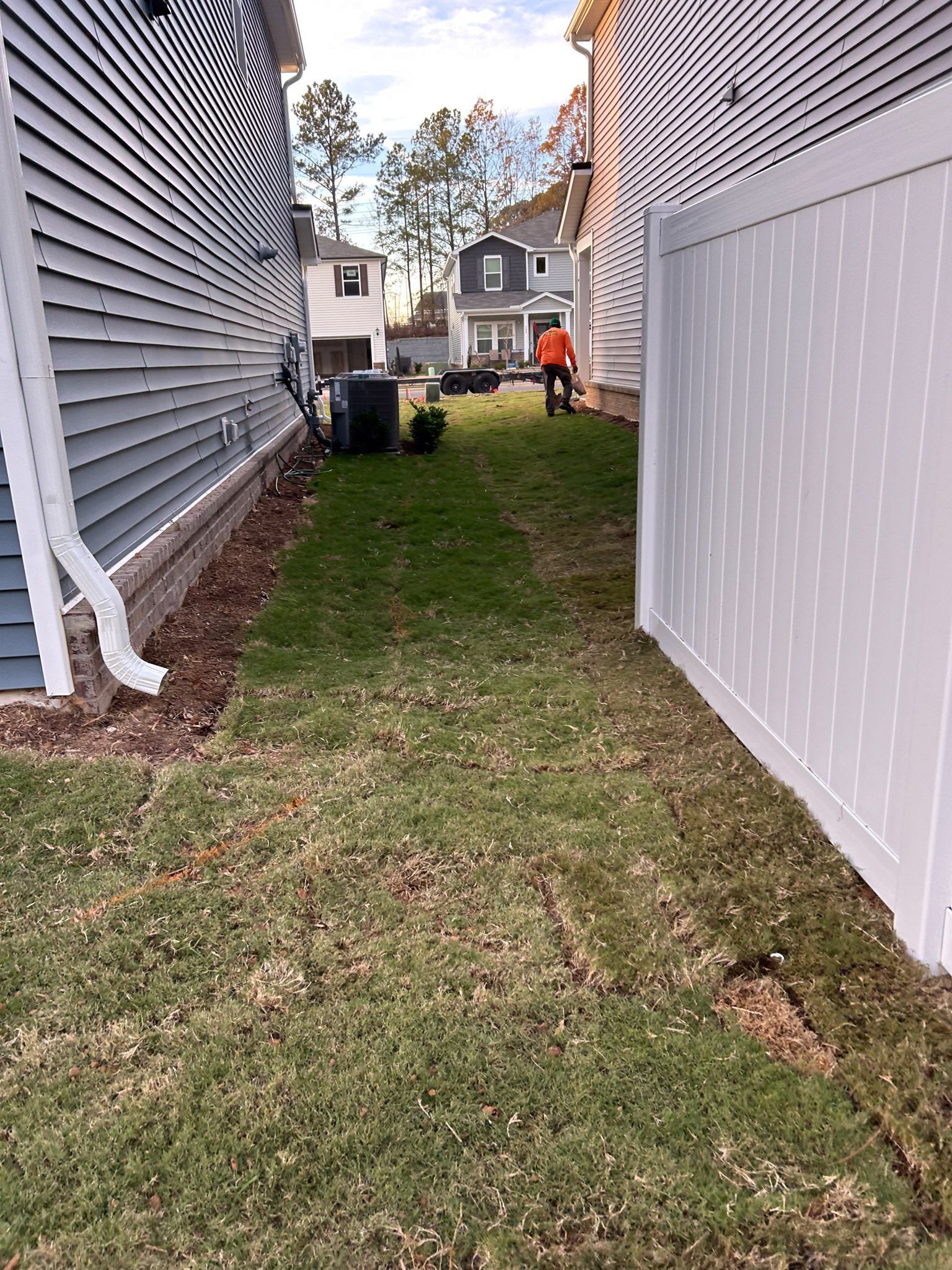 Narrow grassy yard between two houses and a white fence; a person in the distance.