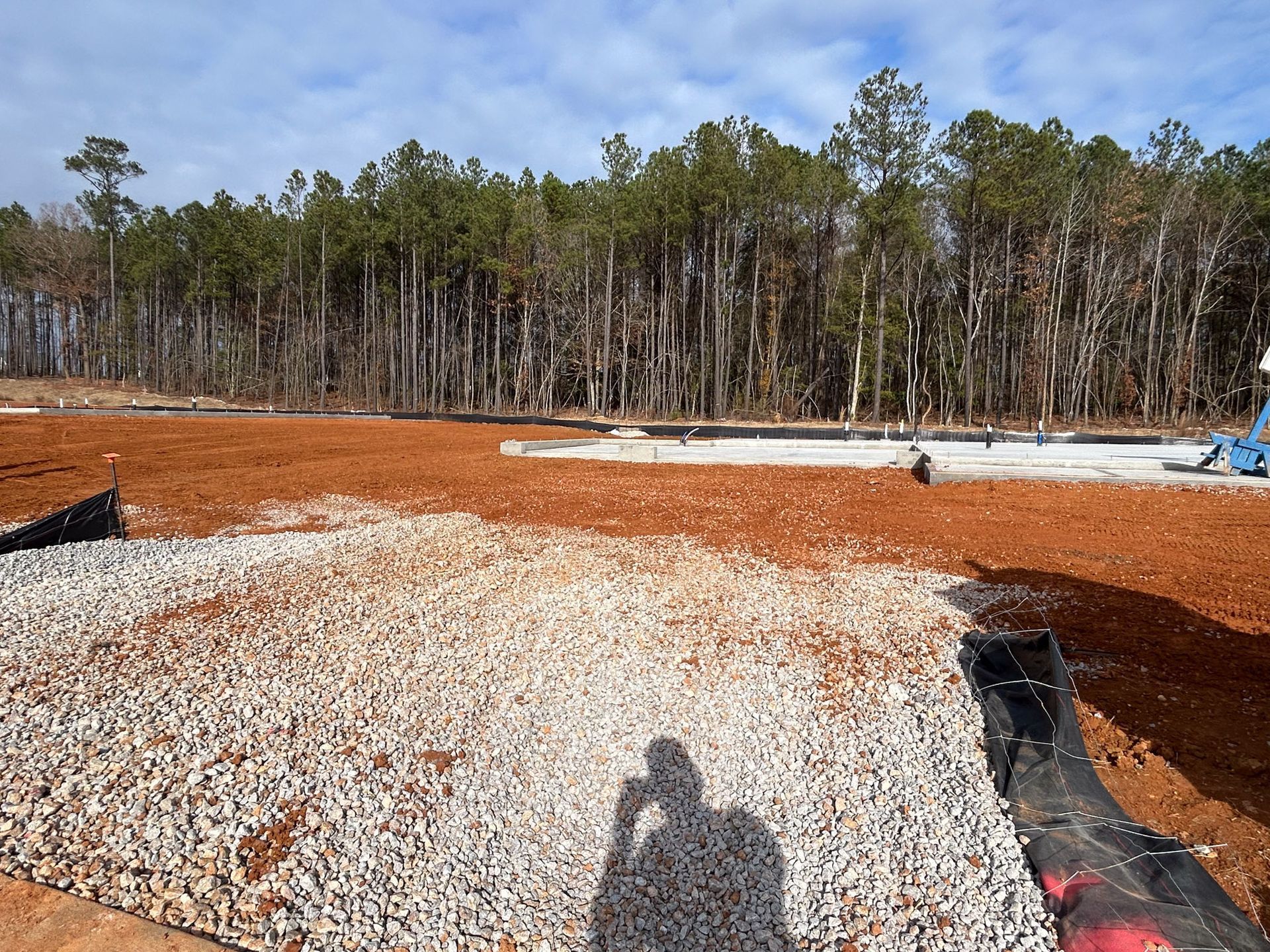 Construction site with gravel and red dirt in foreground, trees in the background, blue sky.