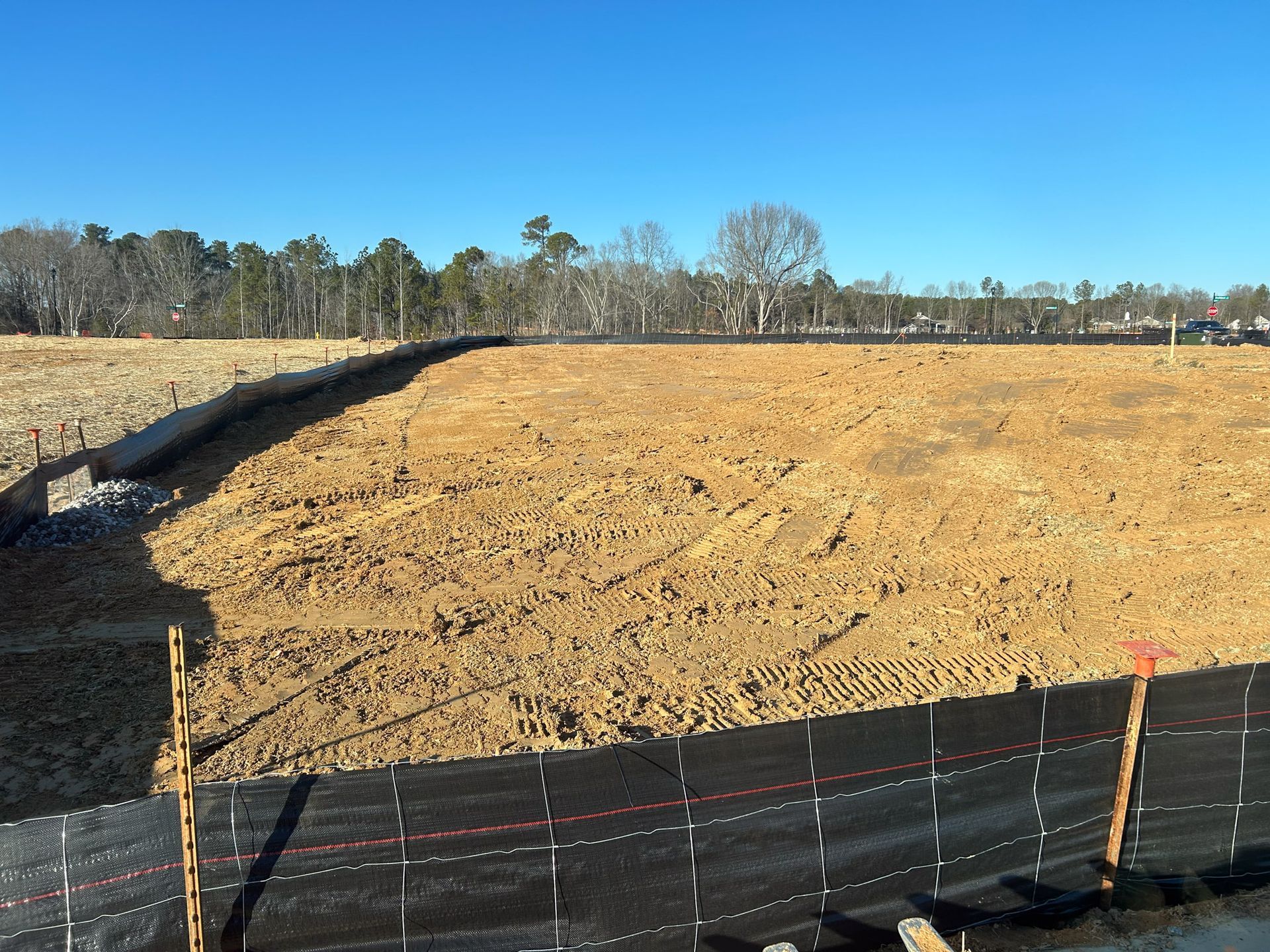 Cleared construction site with dirt, black barrier, and trees under a blue sky.