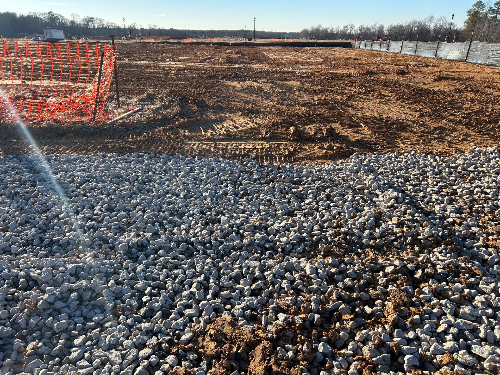 Gravel foreground, dirt construction site, orange safety fencing, clear sky.