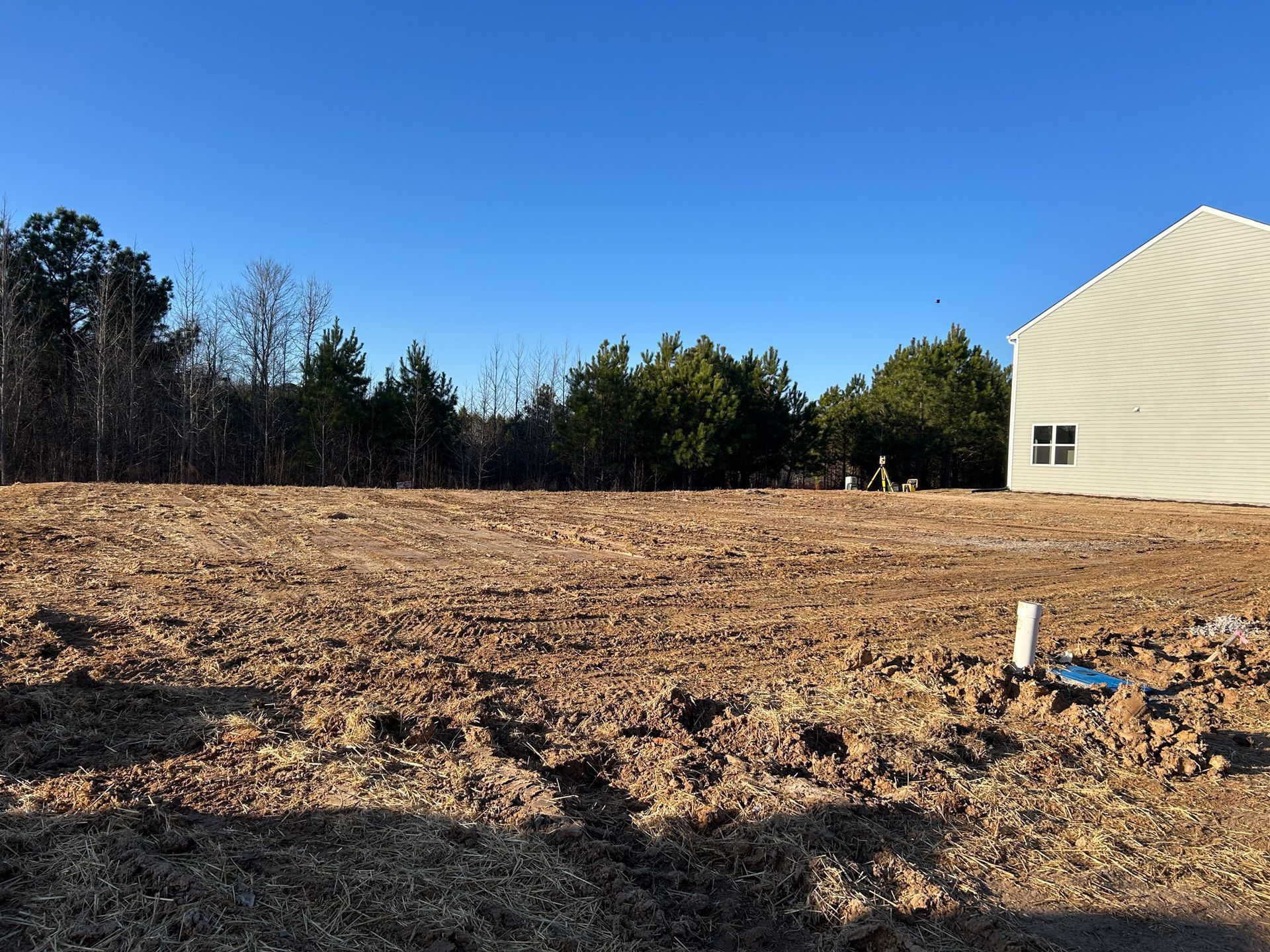 Cleared lot with exposed earth, partially built house on the right, trees in the background, blue sky.