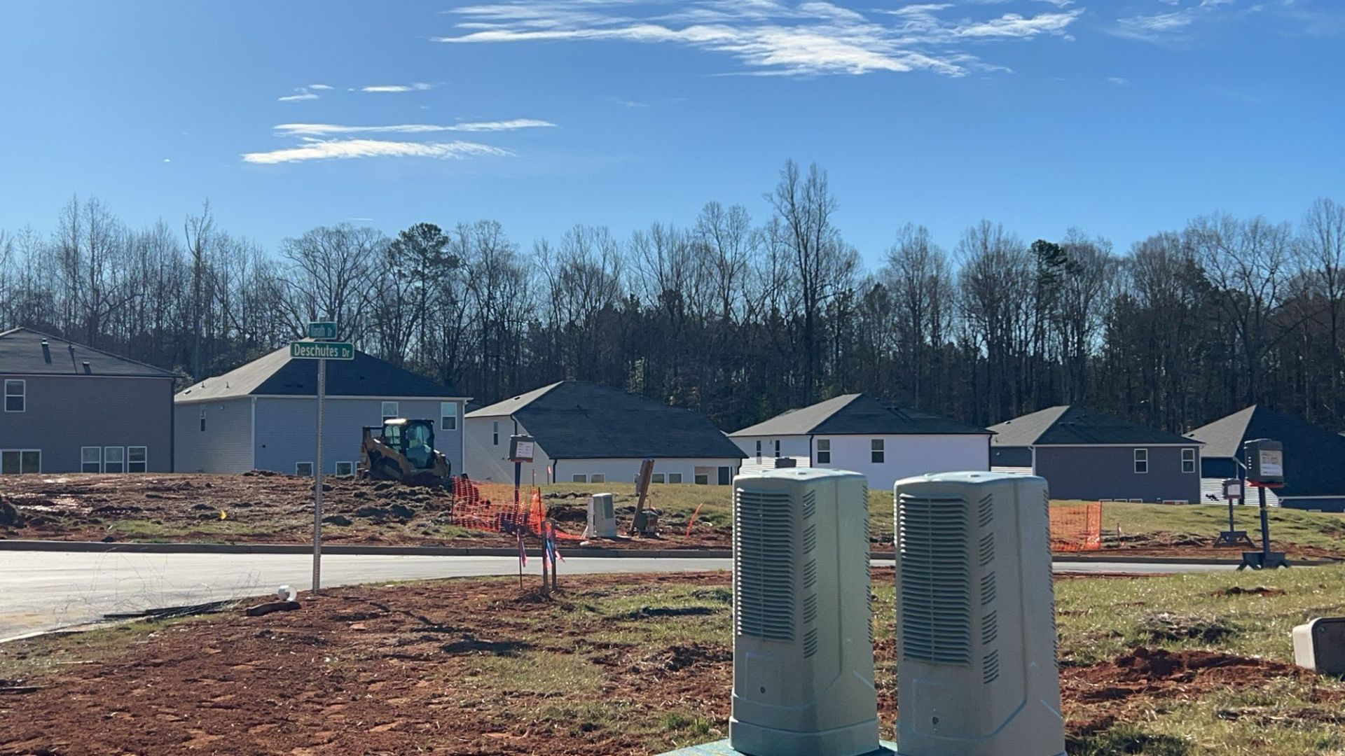 New homes under construction with an excavator in the distance, set against a backdrop of trees and a blue sky.