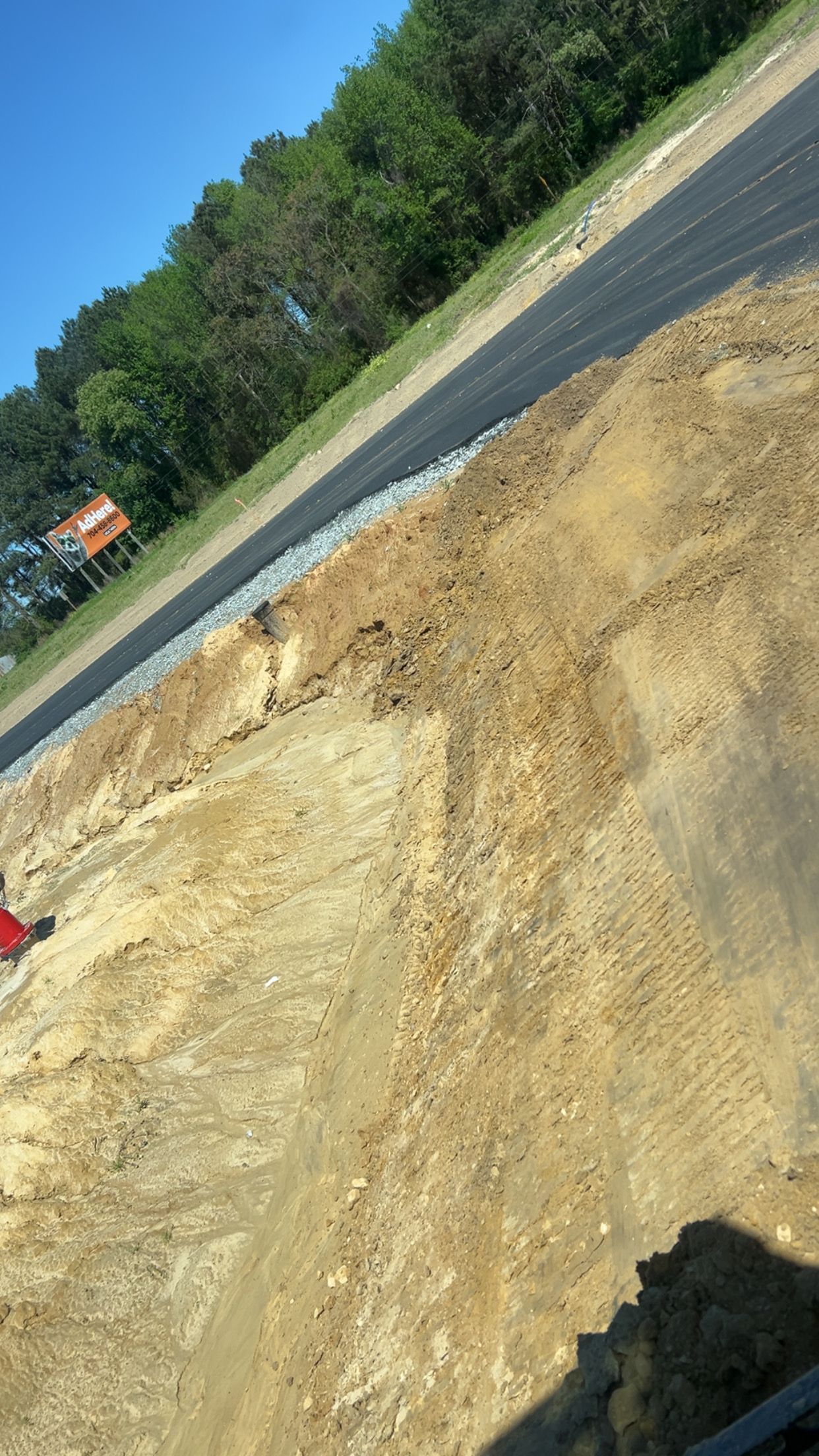 Dirt embankment next to new road; trees in the background under a blue sky.