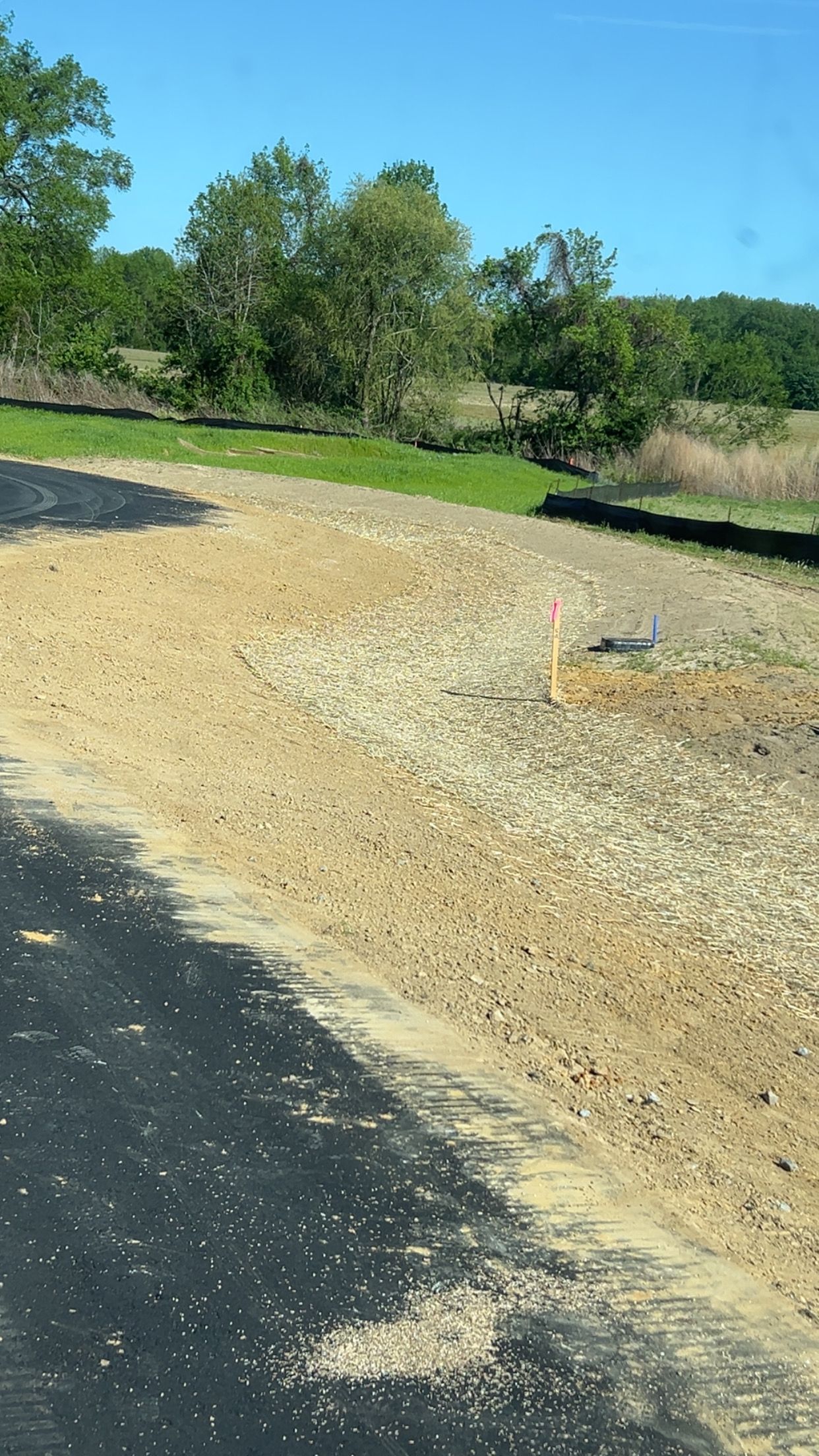 Asphalt road edge next to gravel ground, leading to a grassy area with trees and a blue sky.