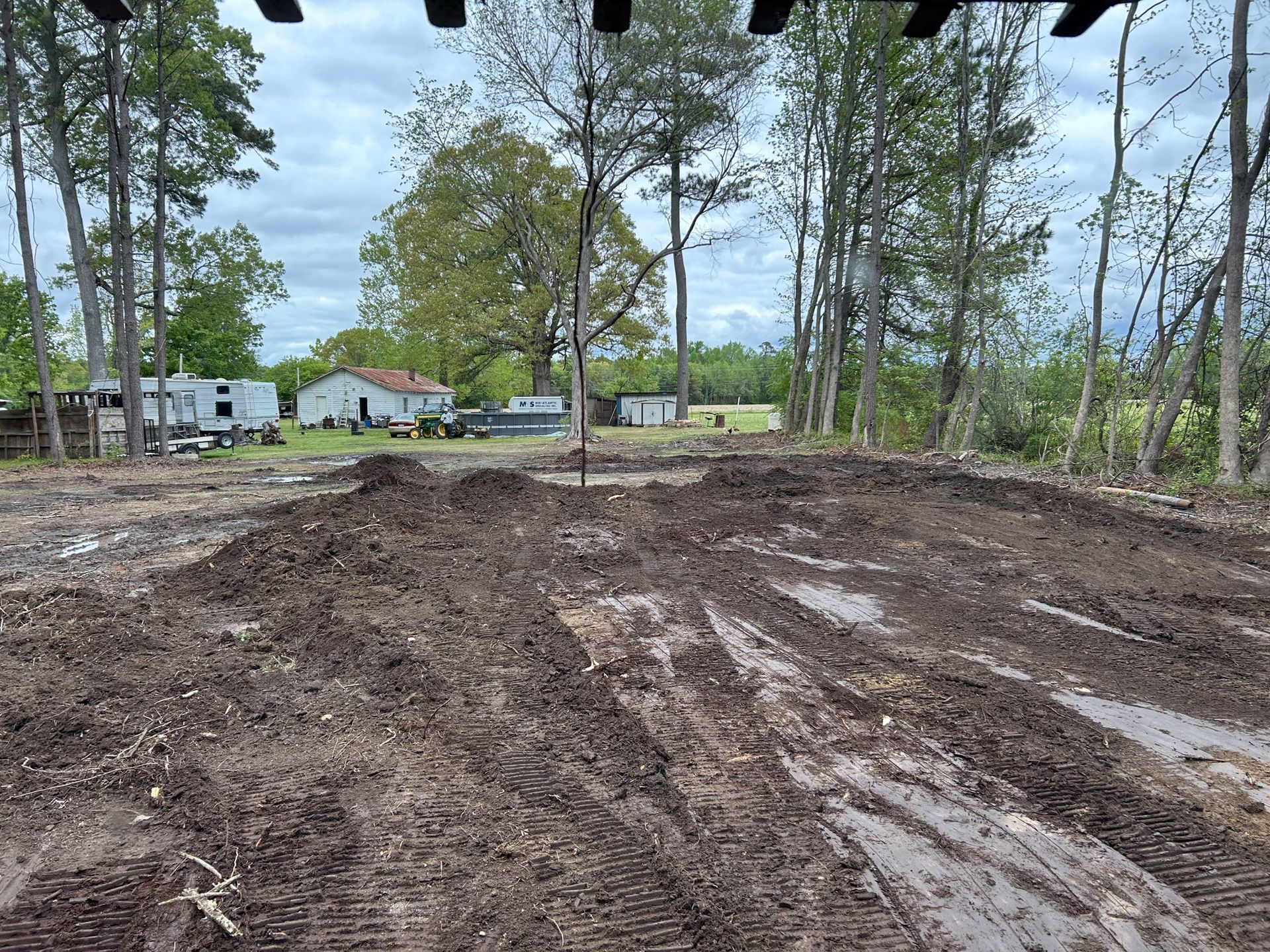 Muddy construction site with piles of dirt, trees, and houses in the background under a cloudy sky.