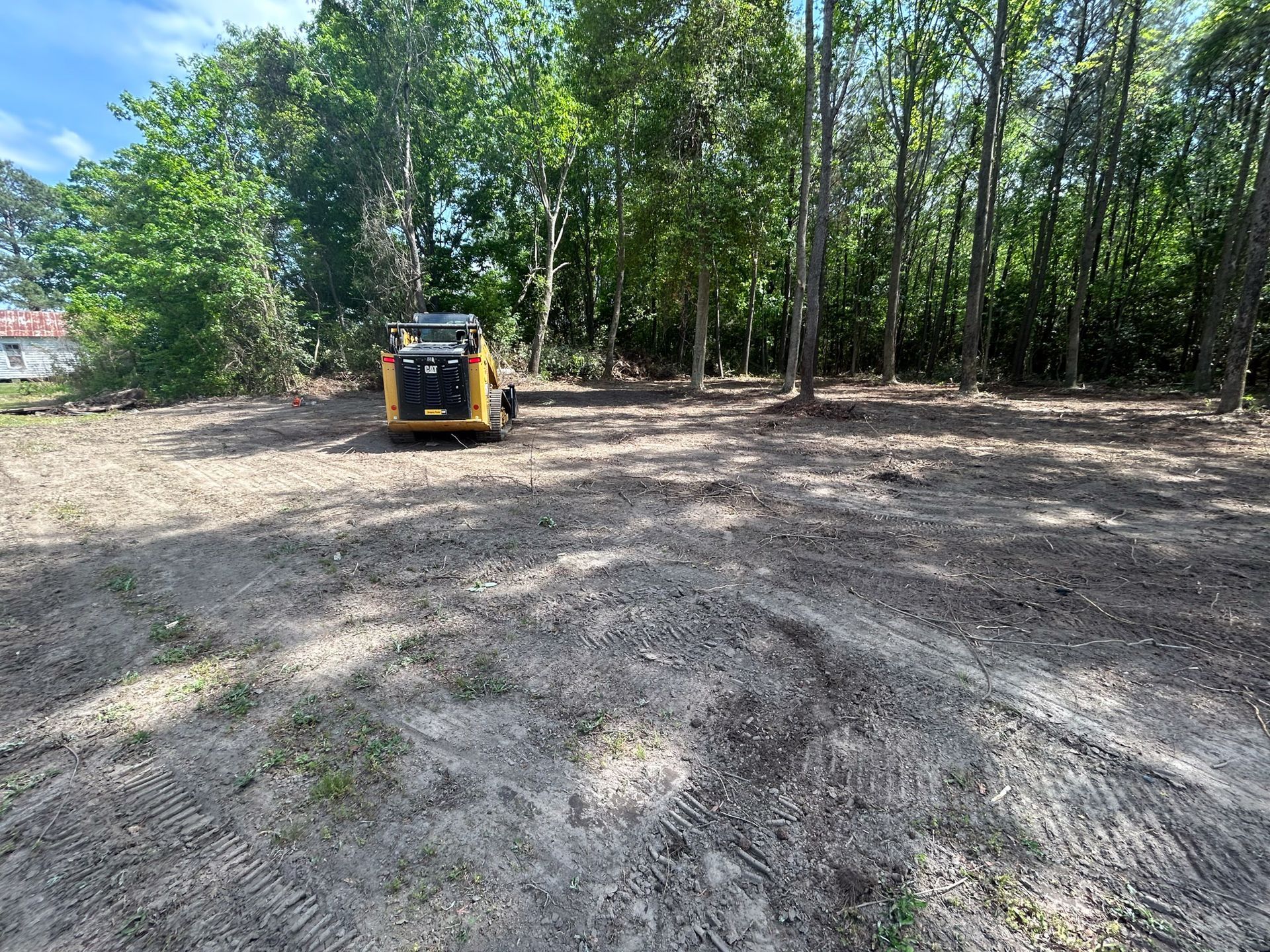 A cleared dirt lot with a small yellow construction vehicle, trees in the background, blue sky.