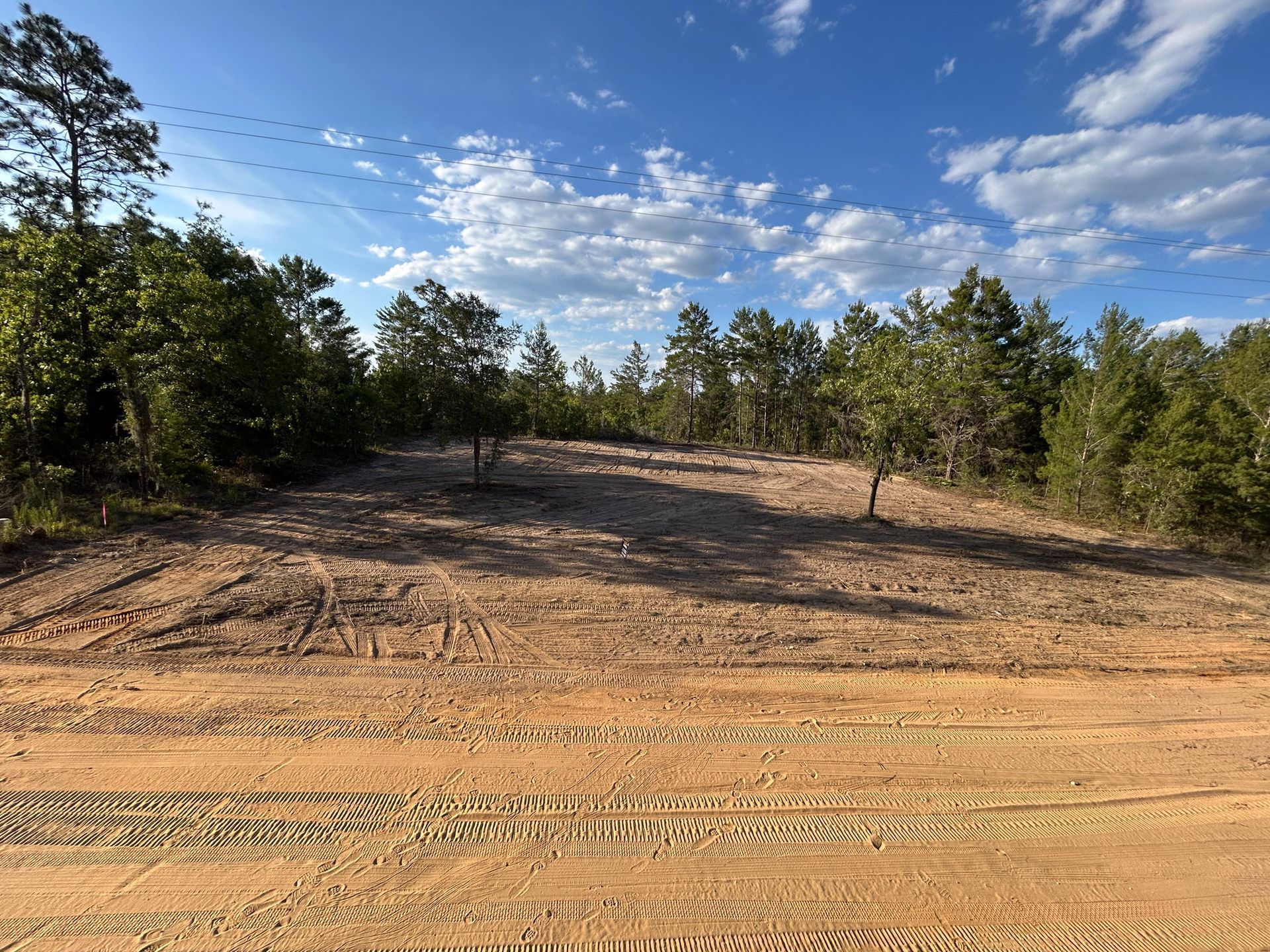 Cleared dirt lot with tire tracks, surrounded by trees under a partly cloudy blue sky.