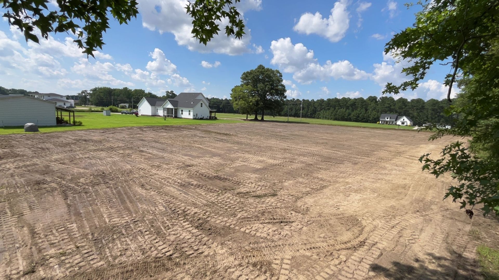 Cleared, empty lot with visible tire tracks, houses in the background, blue sky with clouds.
