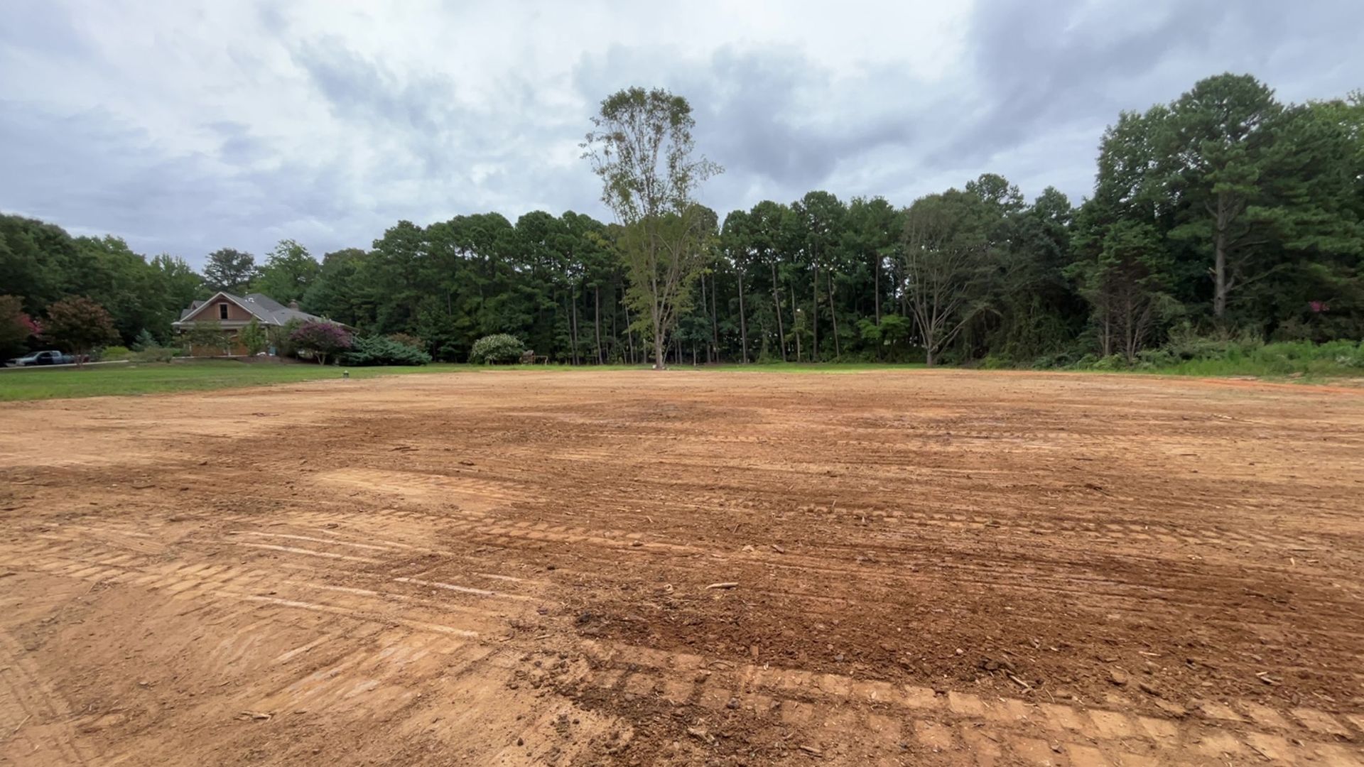 Cleared, brown dirt field with tire tracks, trees, and a house under a cloudy sky.