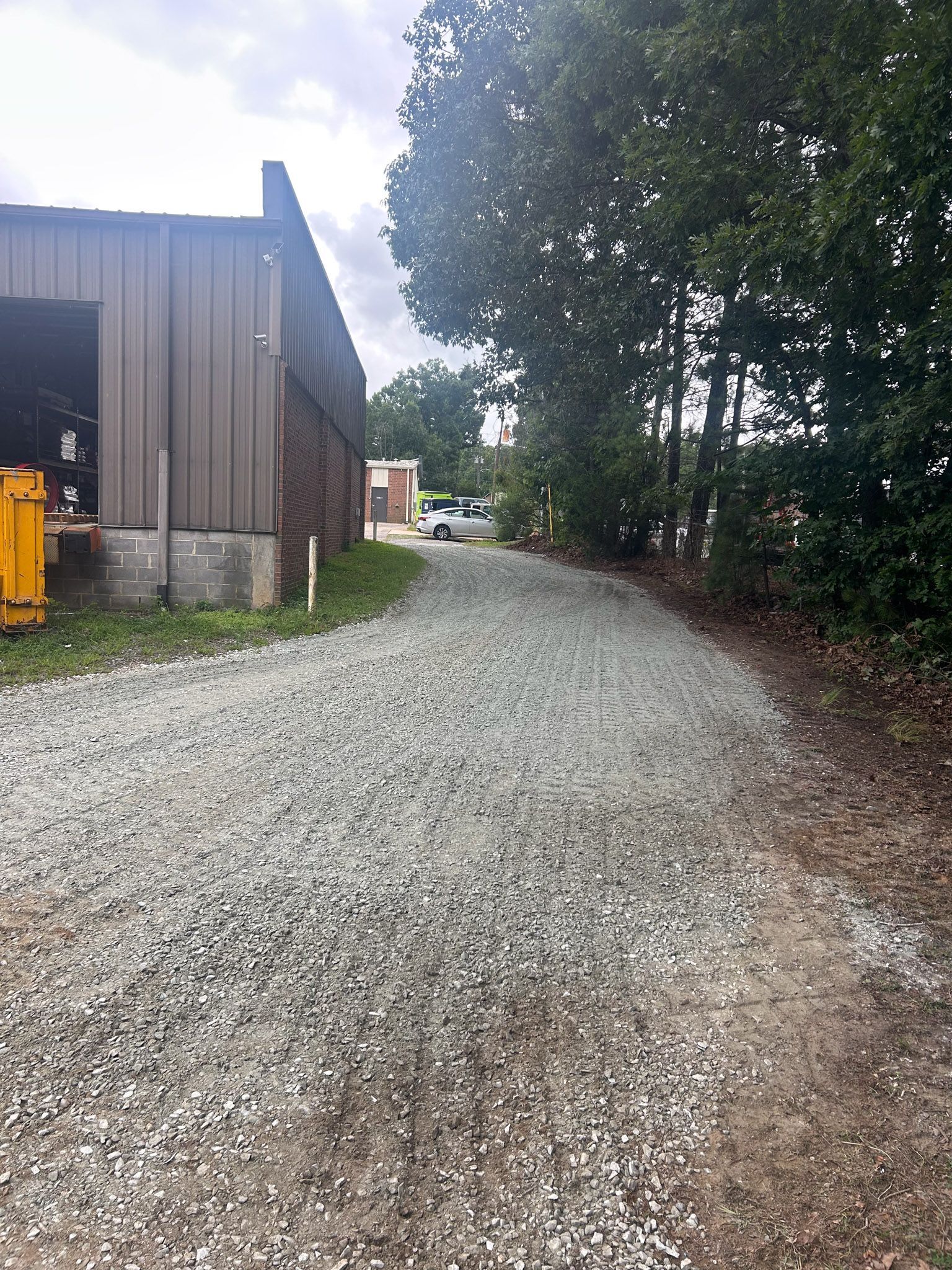 Gravel driveway next to a building and trees, leading to a street with a car.