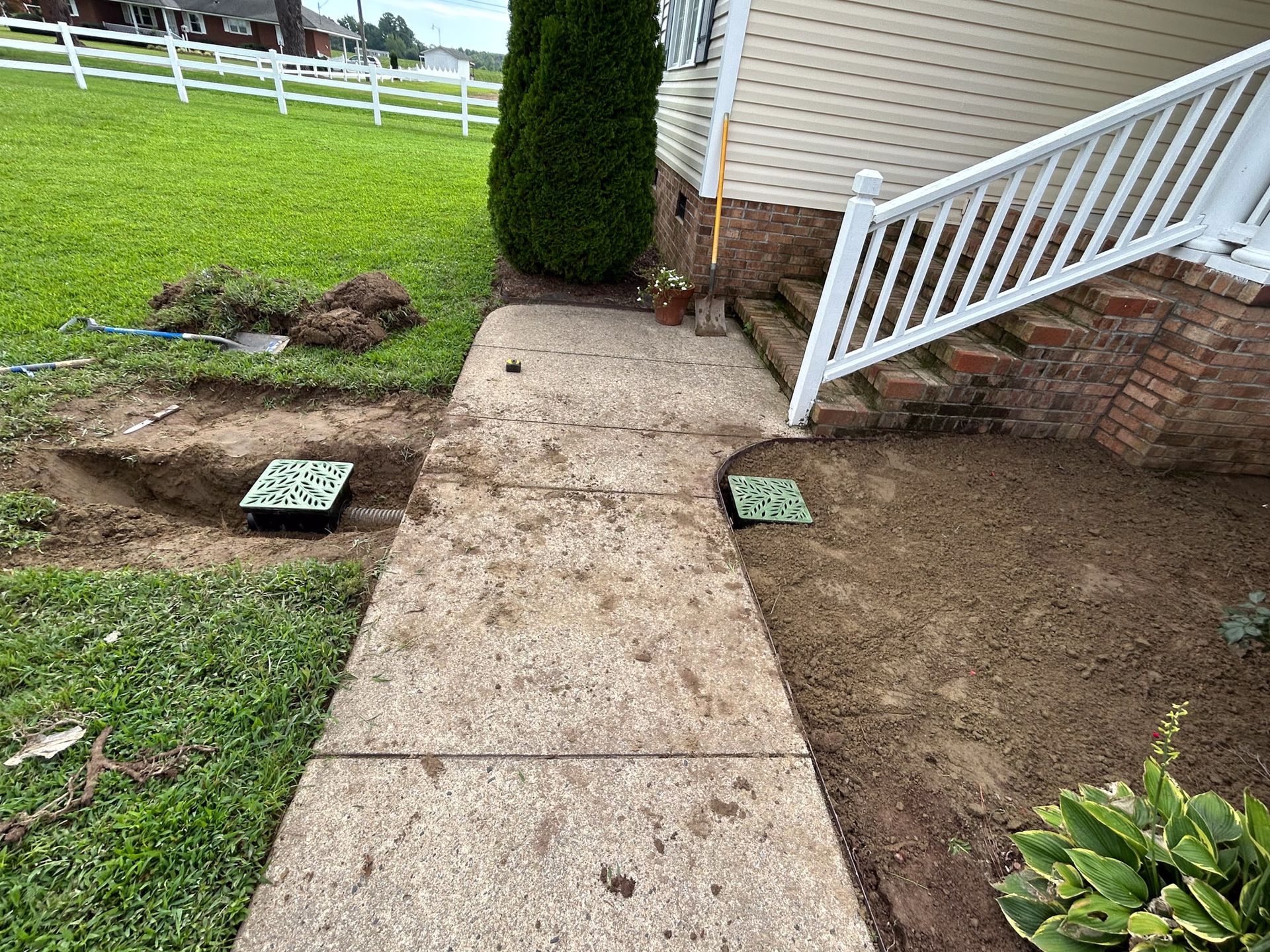Sidewalk with two installed drainage grates and a trench beside it. Residential setting with stairs.