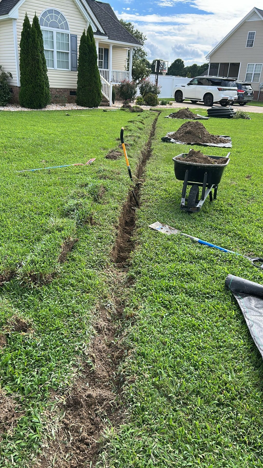 Trench dug in a grassy yard, with tools and a wheelbarrow nearby. House in background.