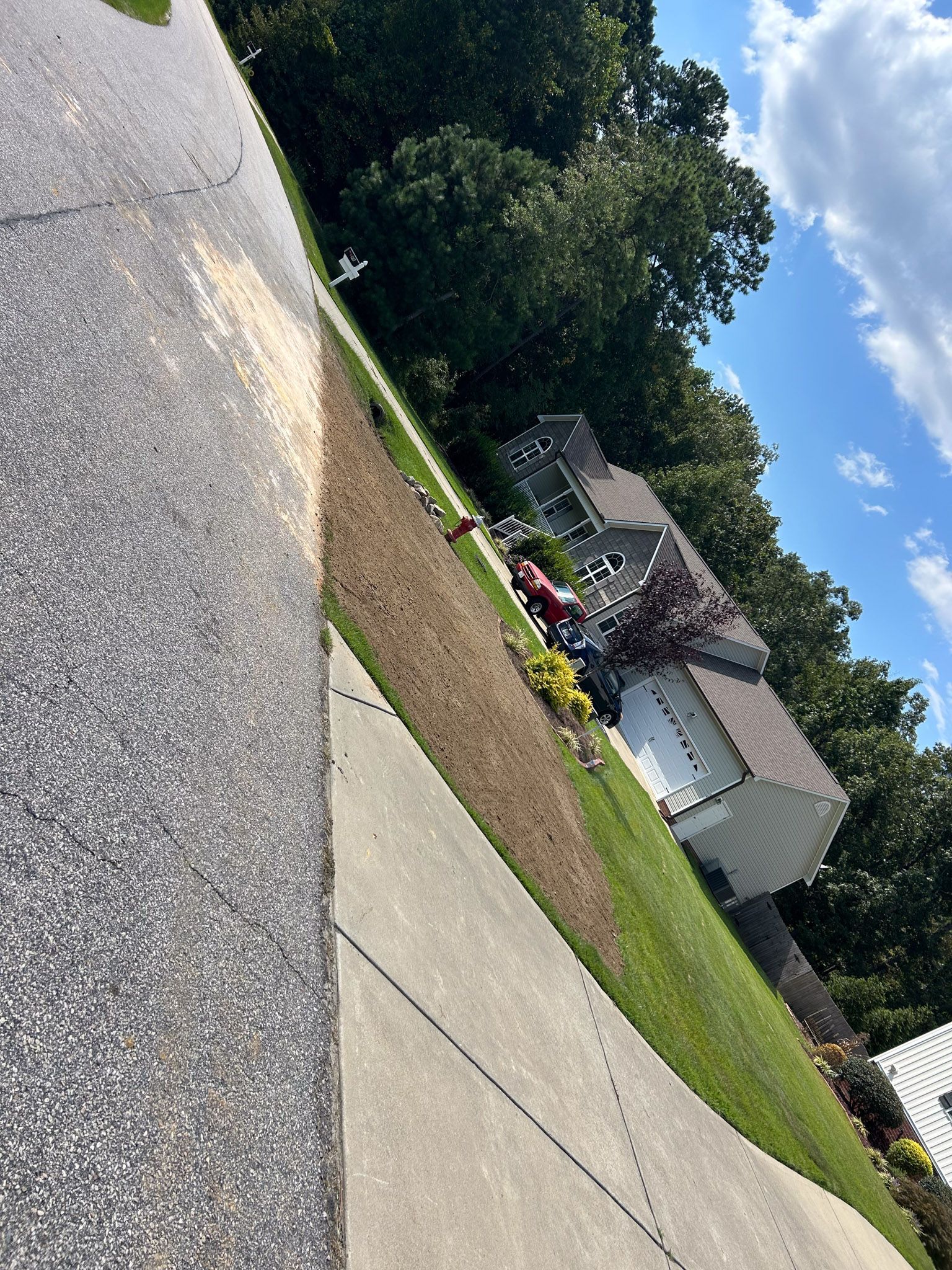 Asphalt road and sidewalk beside a green, sloped lawn leading to a house under a partly cloudy blue sky.