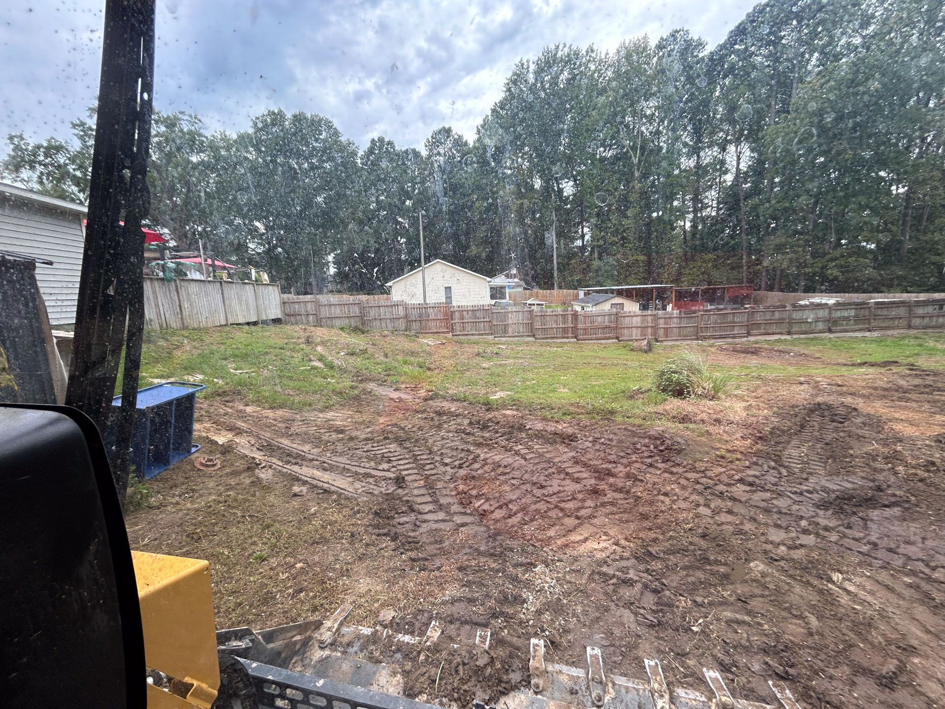 A muddy construction site viewed from a yellow excavator with a house in the background.