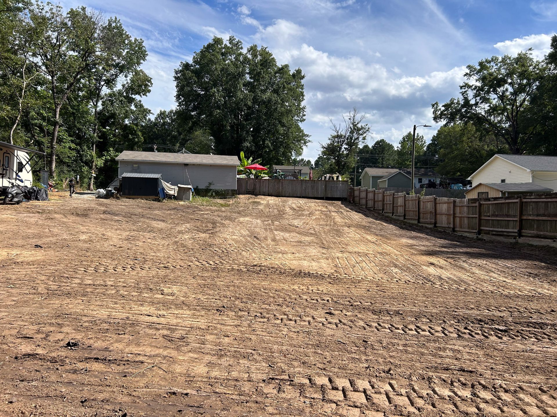 Cleared dirt lot with a wooden fence bordering the edges, surrounded by trees and buildings under a cloudy sky.