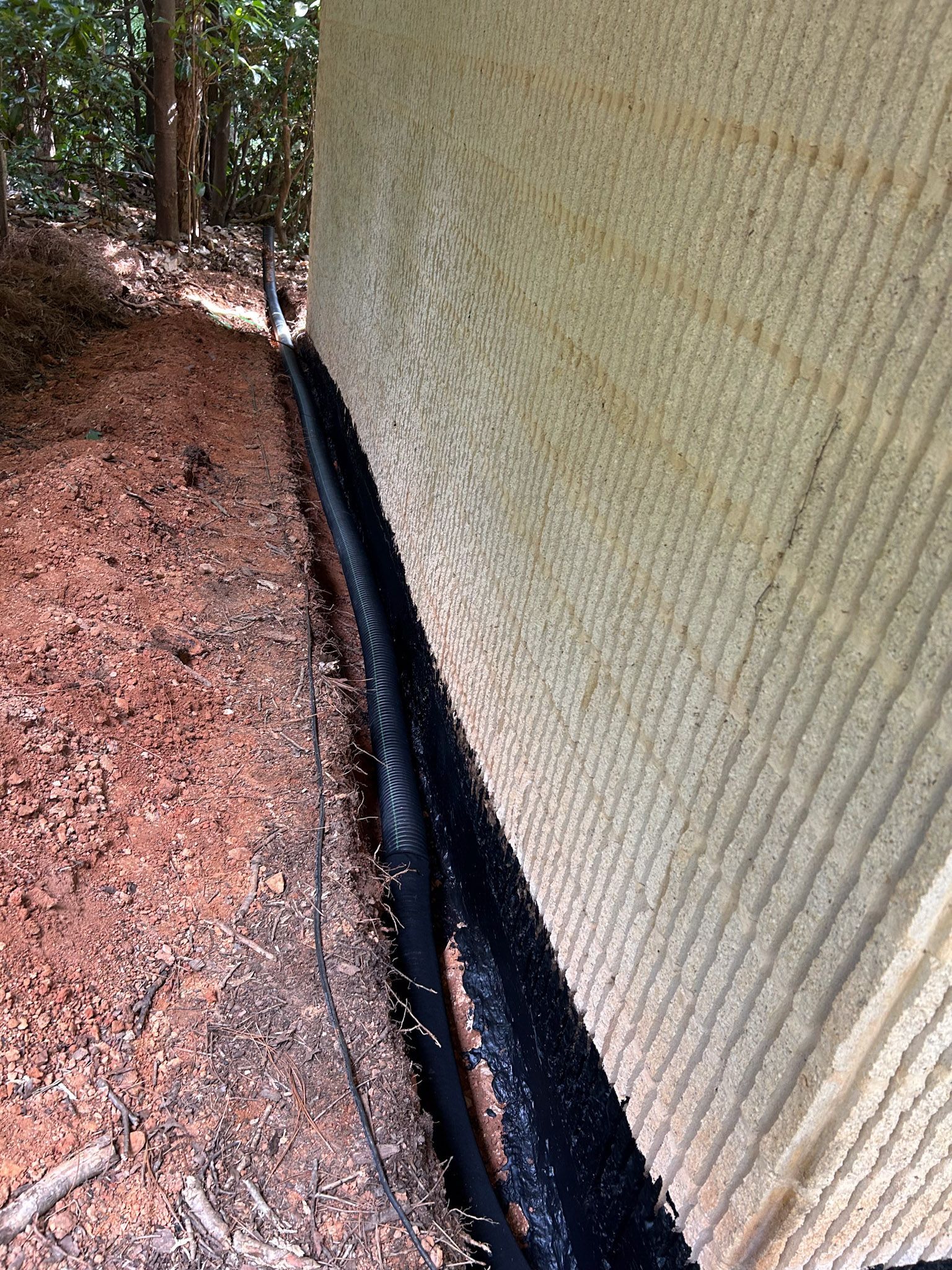 A textured light beige wall next to a dark-colored drainage system on a dirt path, outdoors.