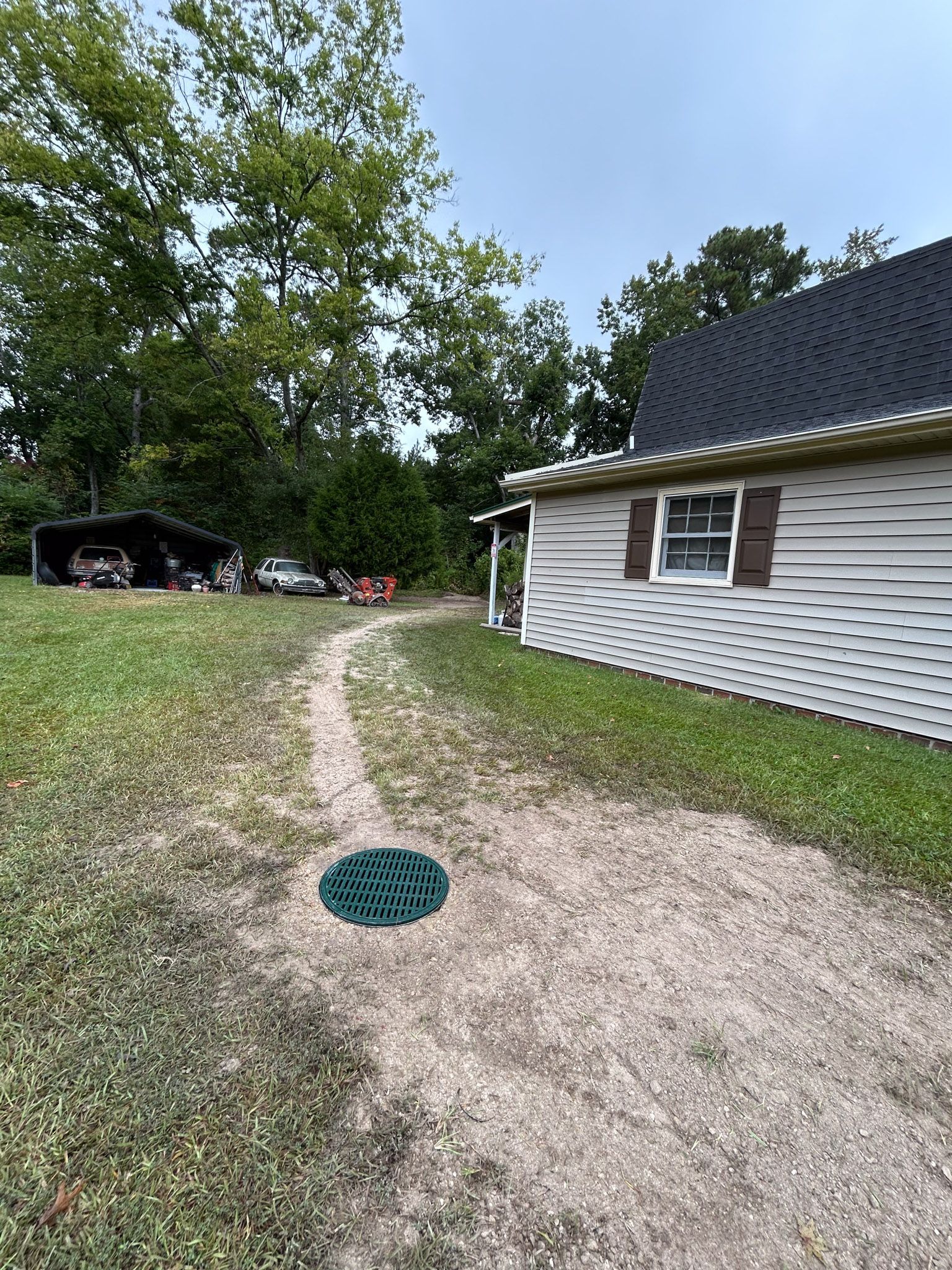 Gravel driveway leads to a shed; green septic lid in foreground. Trees and overcast sky in background.