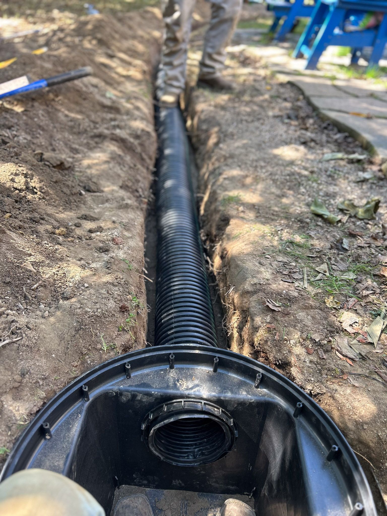 A black drainage pipe in a trench with a person's legs visible in the background, near a black catch basin.
