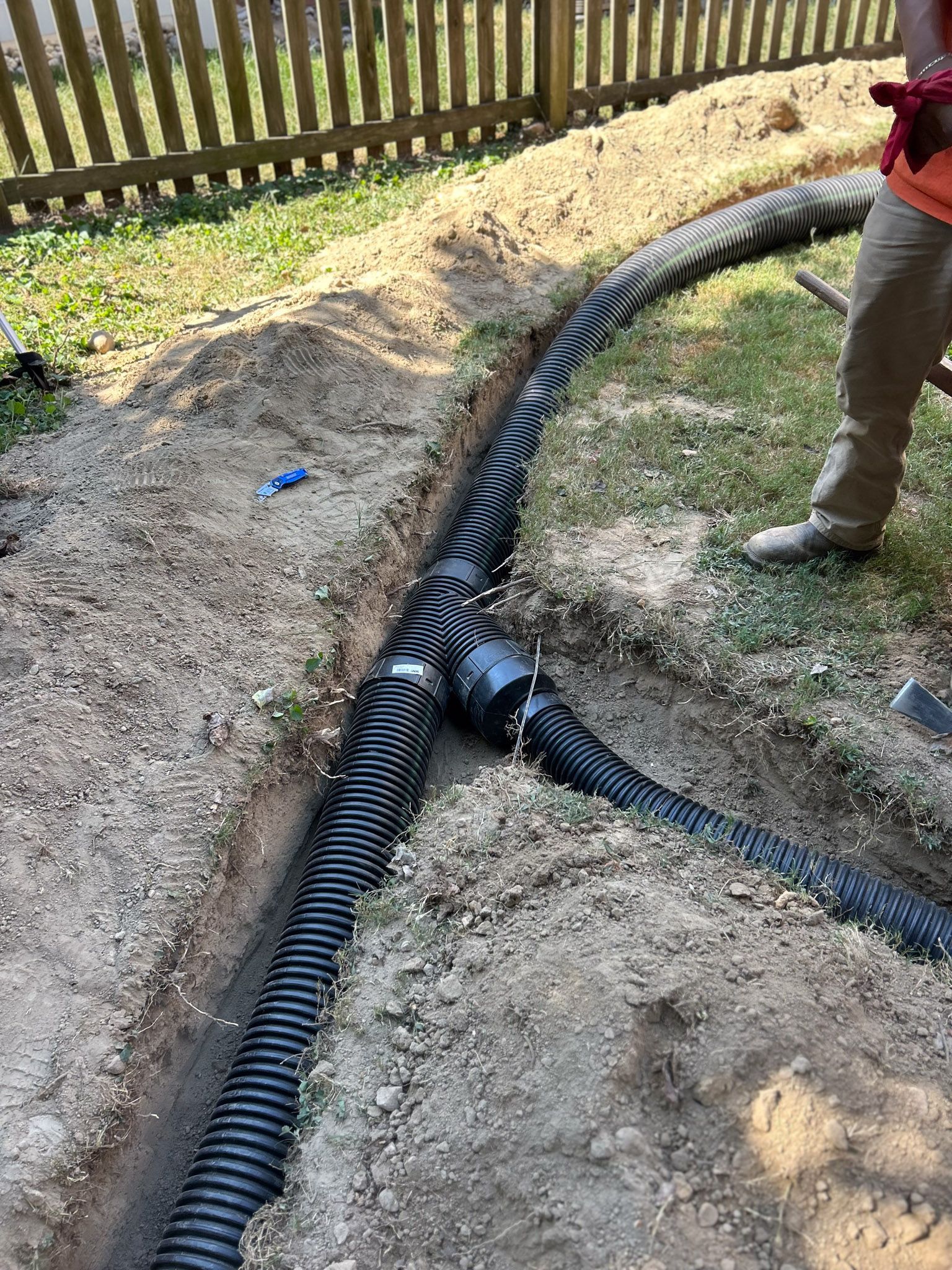 A trench with black corrugated drain pipe, joined by a fitting, near a fence, with a person standing by.