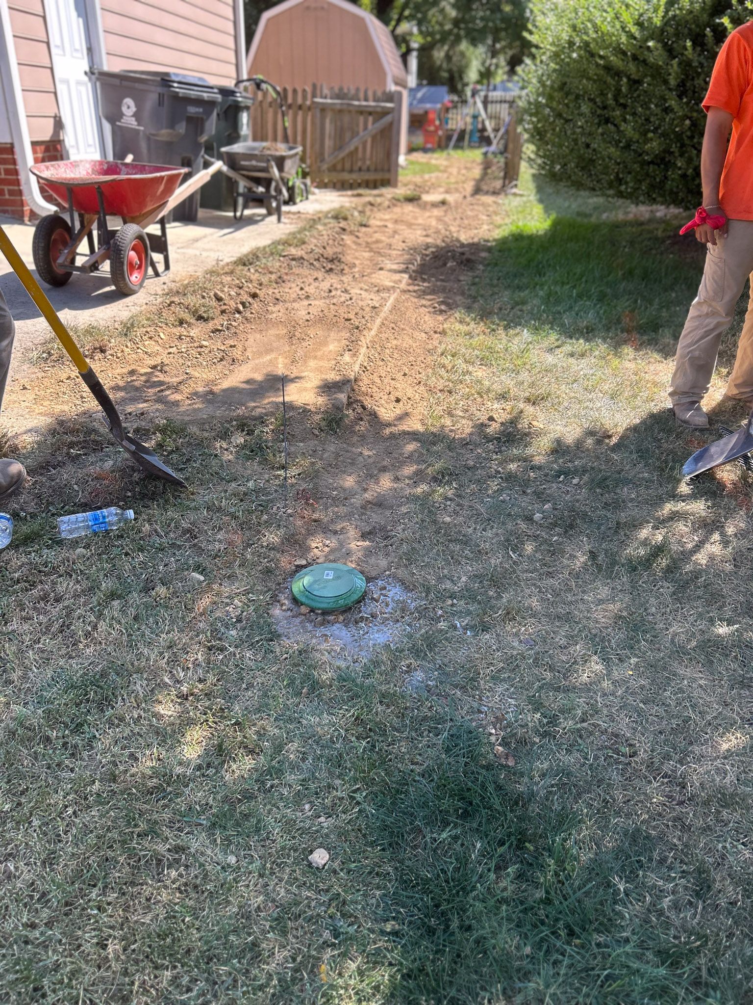 Workers leveling soil in a yard. A green capped object is in front of them surrounded by grass.