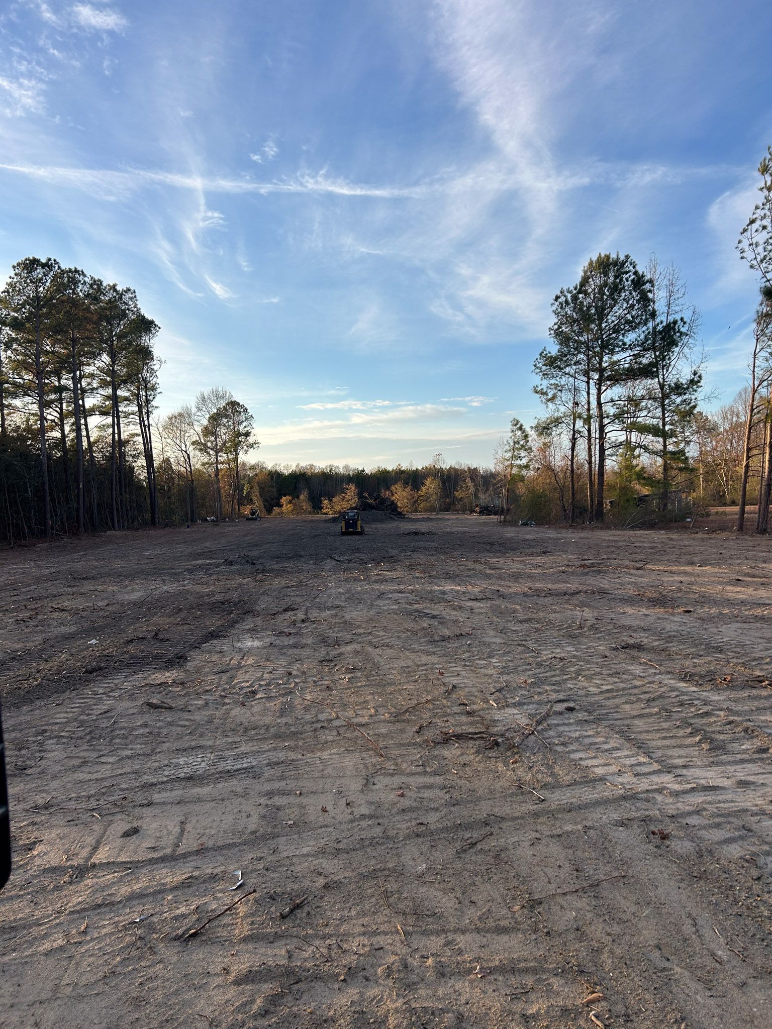 Cleared dirt field with scattered debris, bordered by trees under a partly cloudy sky.