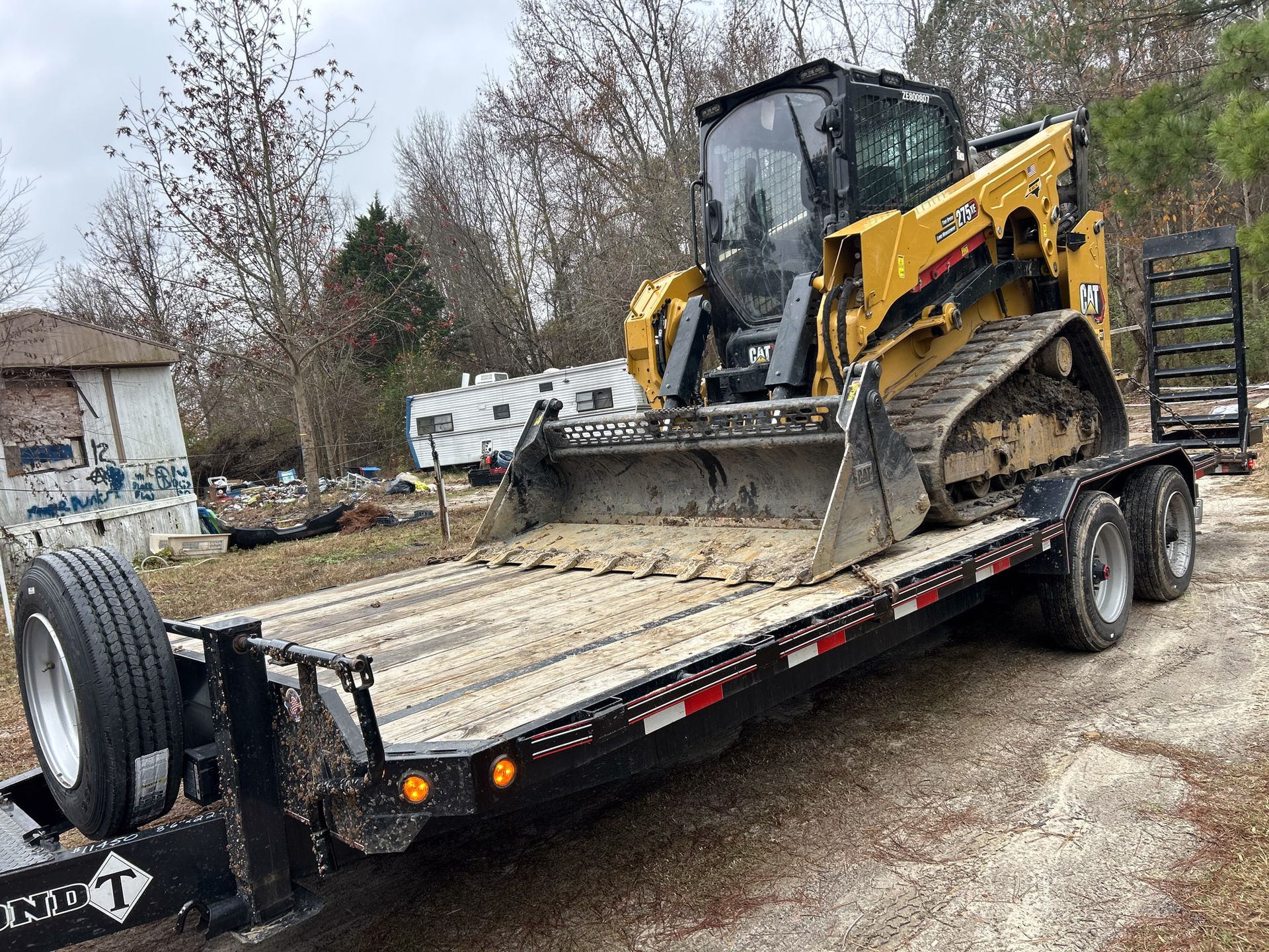 Yellow track loader on a black trailer, parked outside a building.