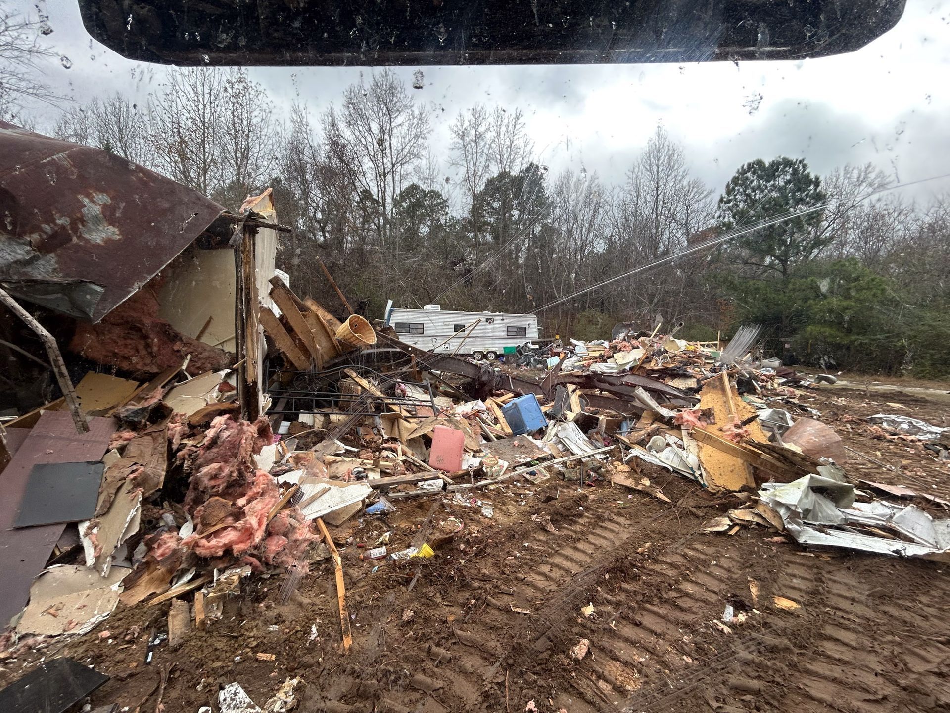 Demolished building debris with a camper in the background, outdoors on a cloudy day.
