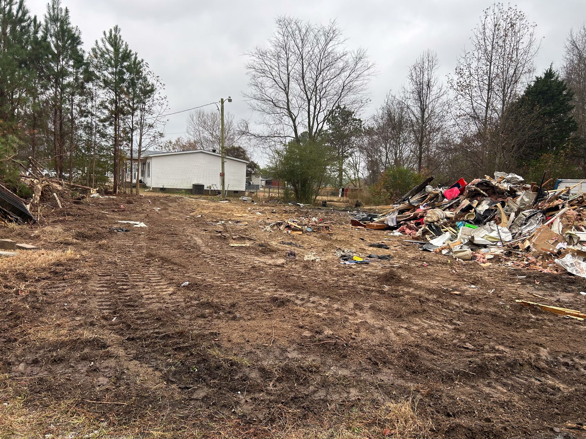 Overgrown dirt lot with a white building in the distance and a large pile of trash on the right side.