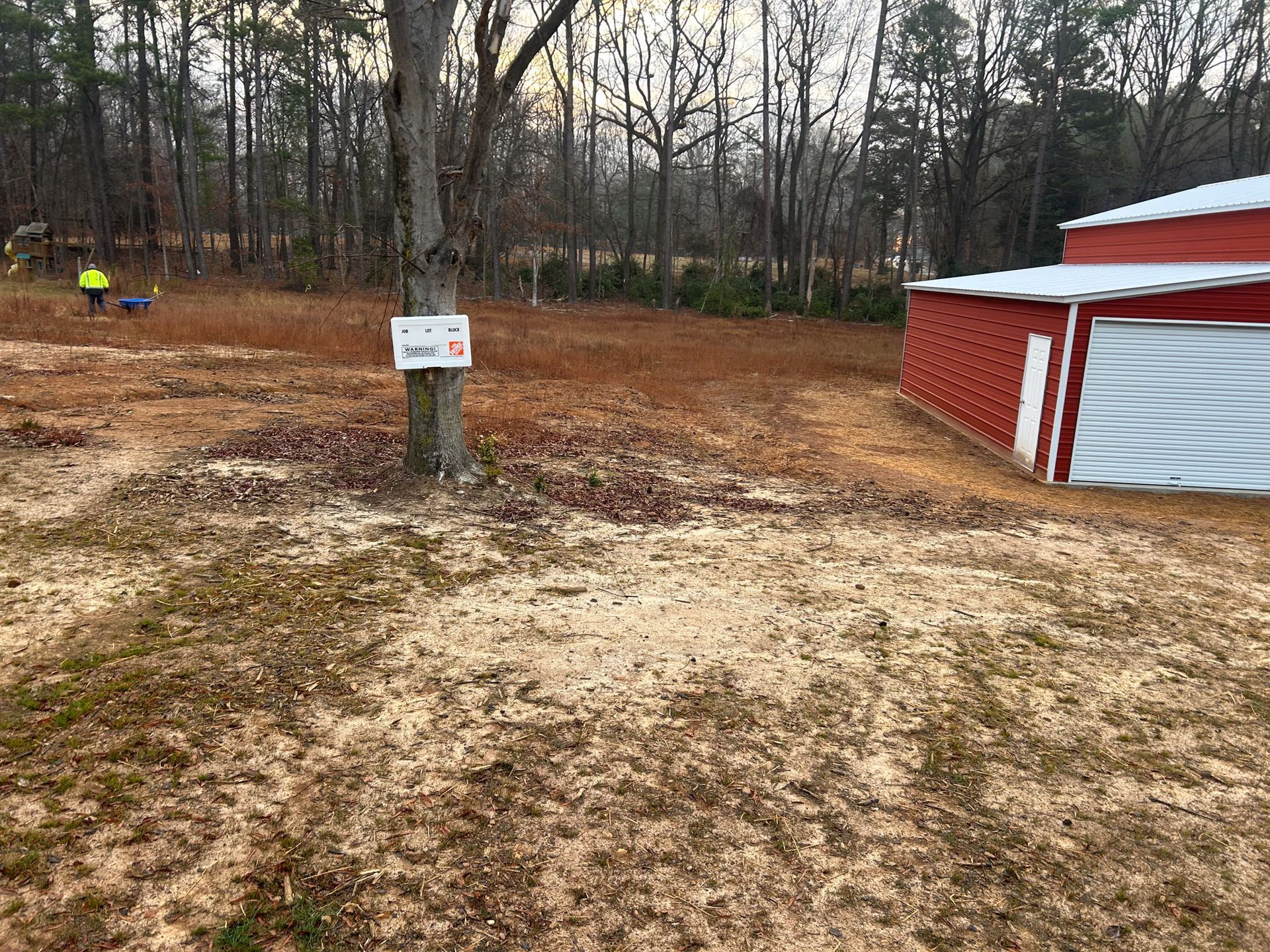 Brown yard with red barn, leafless trees, and person in the distance. Sign on a tree.