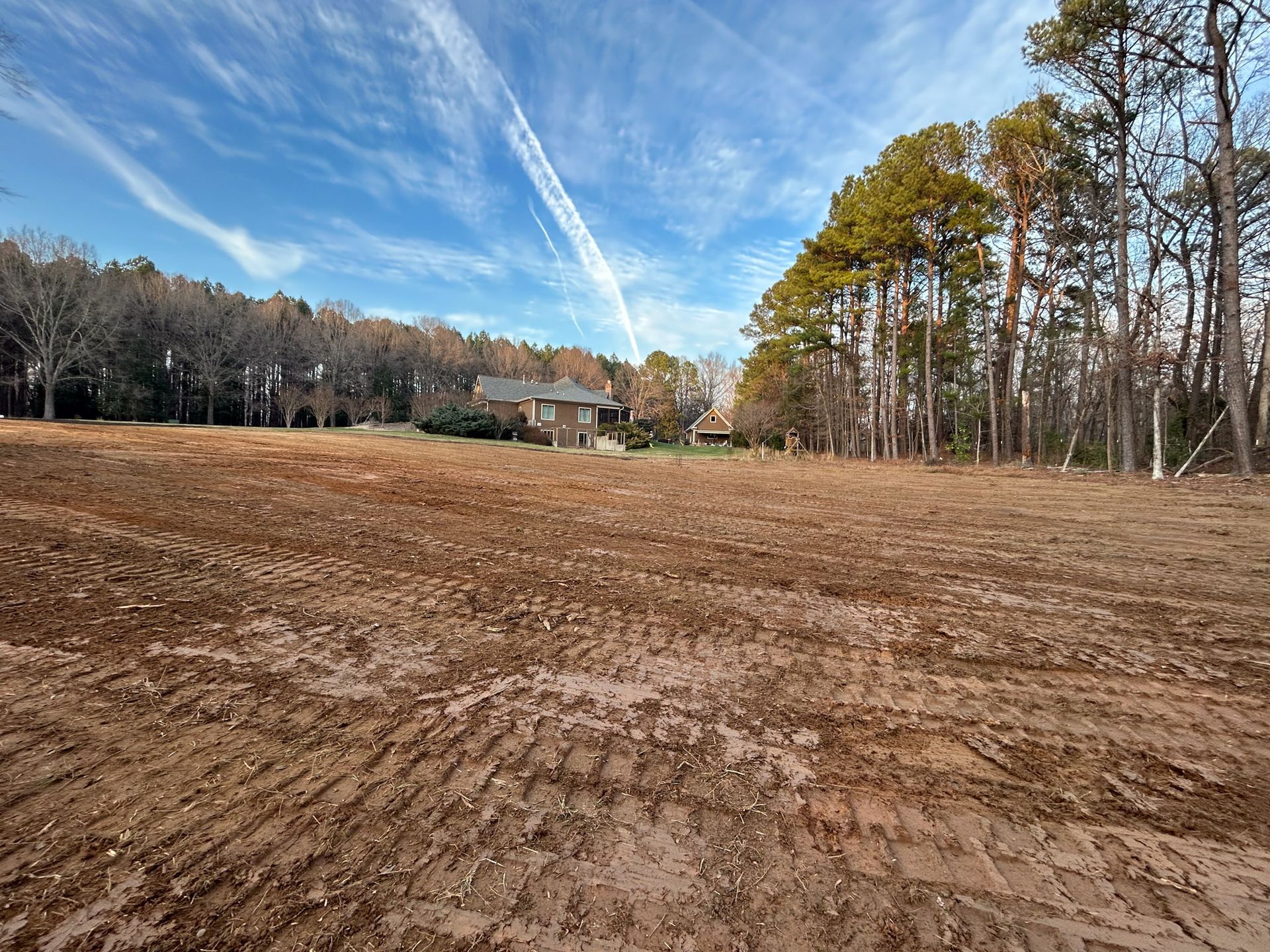 Dirt field with tire tracks, house in the background, trees on either side and a cloudy sky.