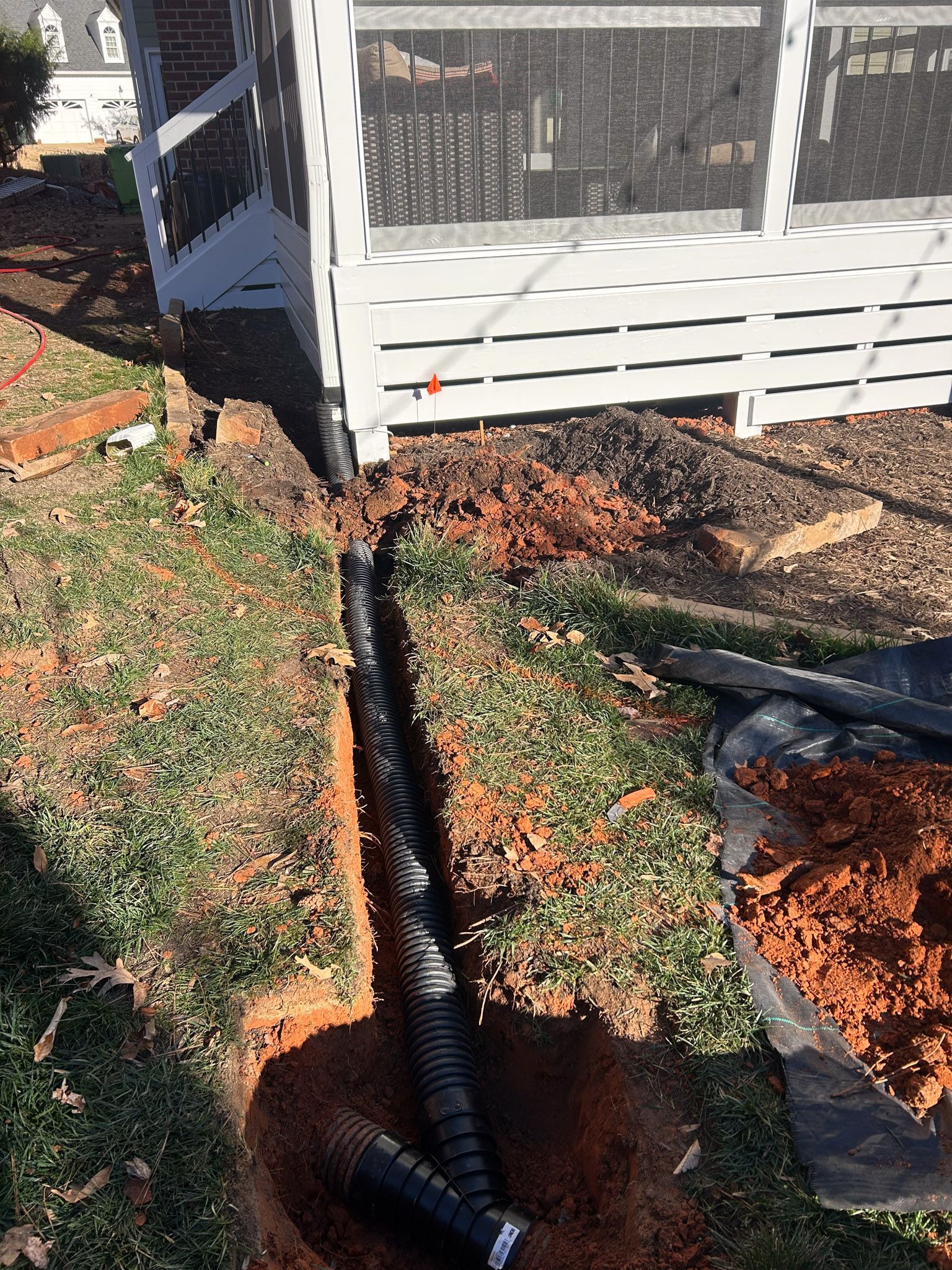 Black drainage pipe in a trench beside a white deck, surrounded by dirt and grass.