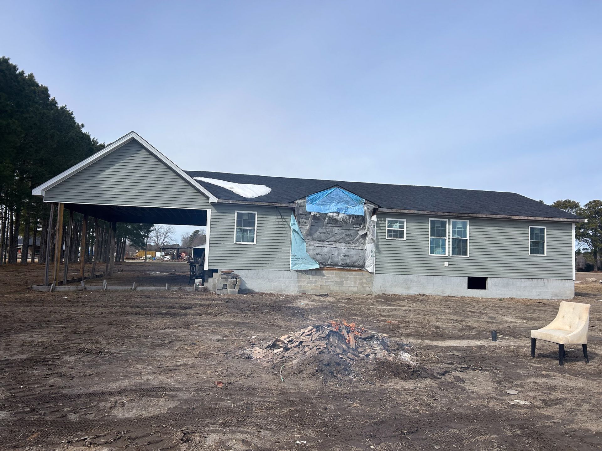 A damaged house with missing siding and a covered carport, in a field, with a pile of debris and chair.
