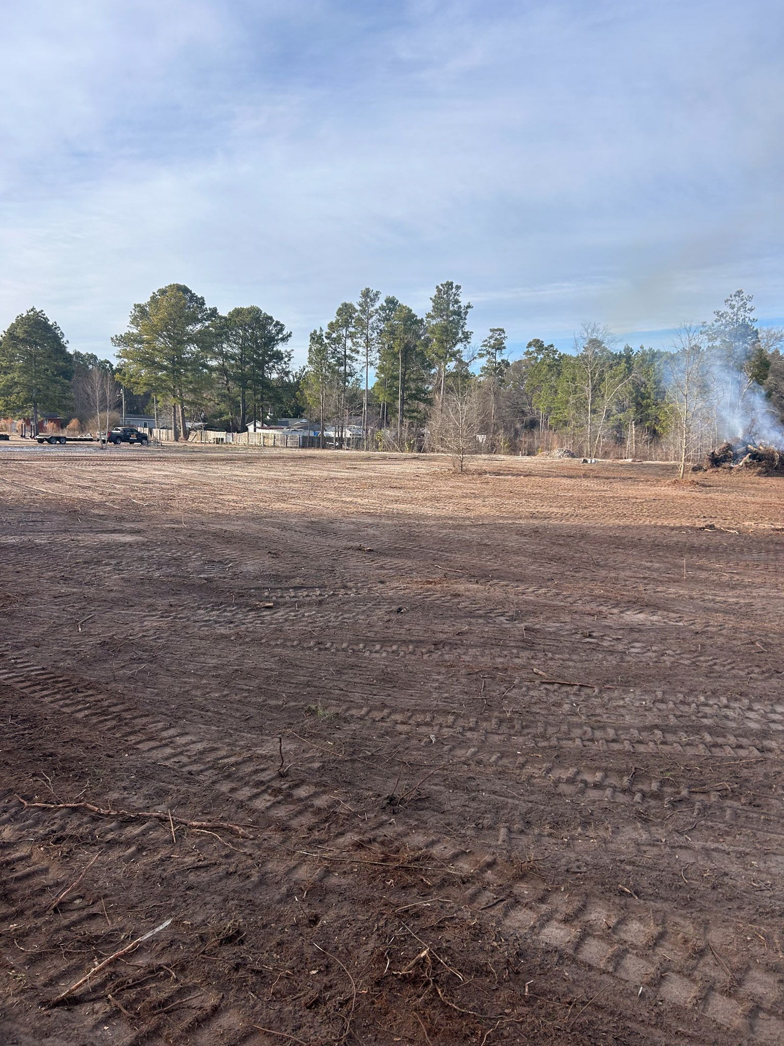 Brown field with tire tracks, trees, and vehicles under a cloudy blue sky.
