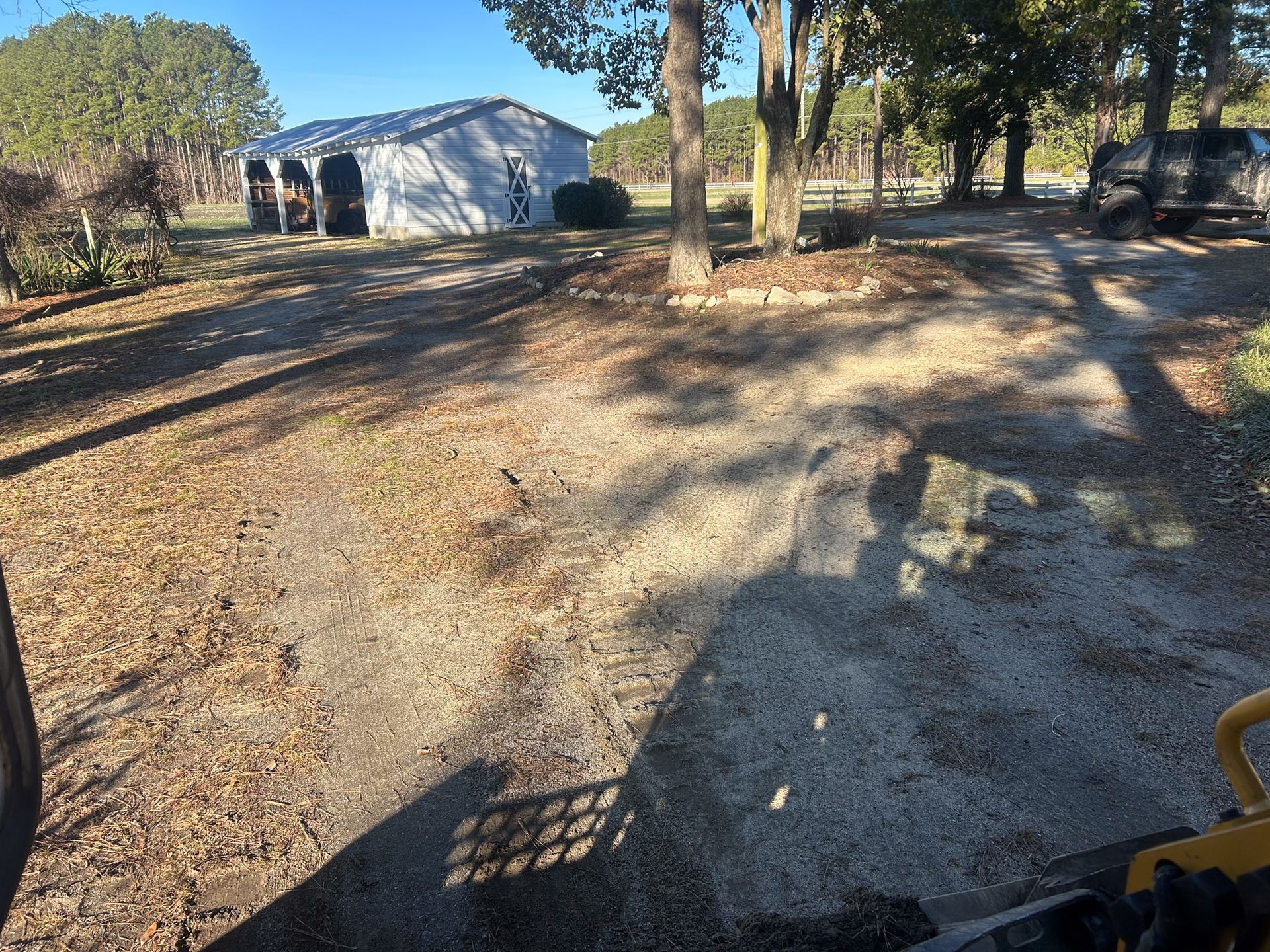 Gravel driveway leading to a white barn on a sunny day. Trees and a dark vehicle are visible.