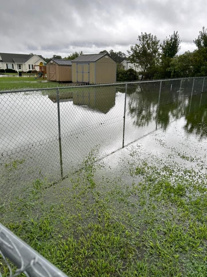 Flooded fenced yard with grass, sheds, and overcast sky.