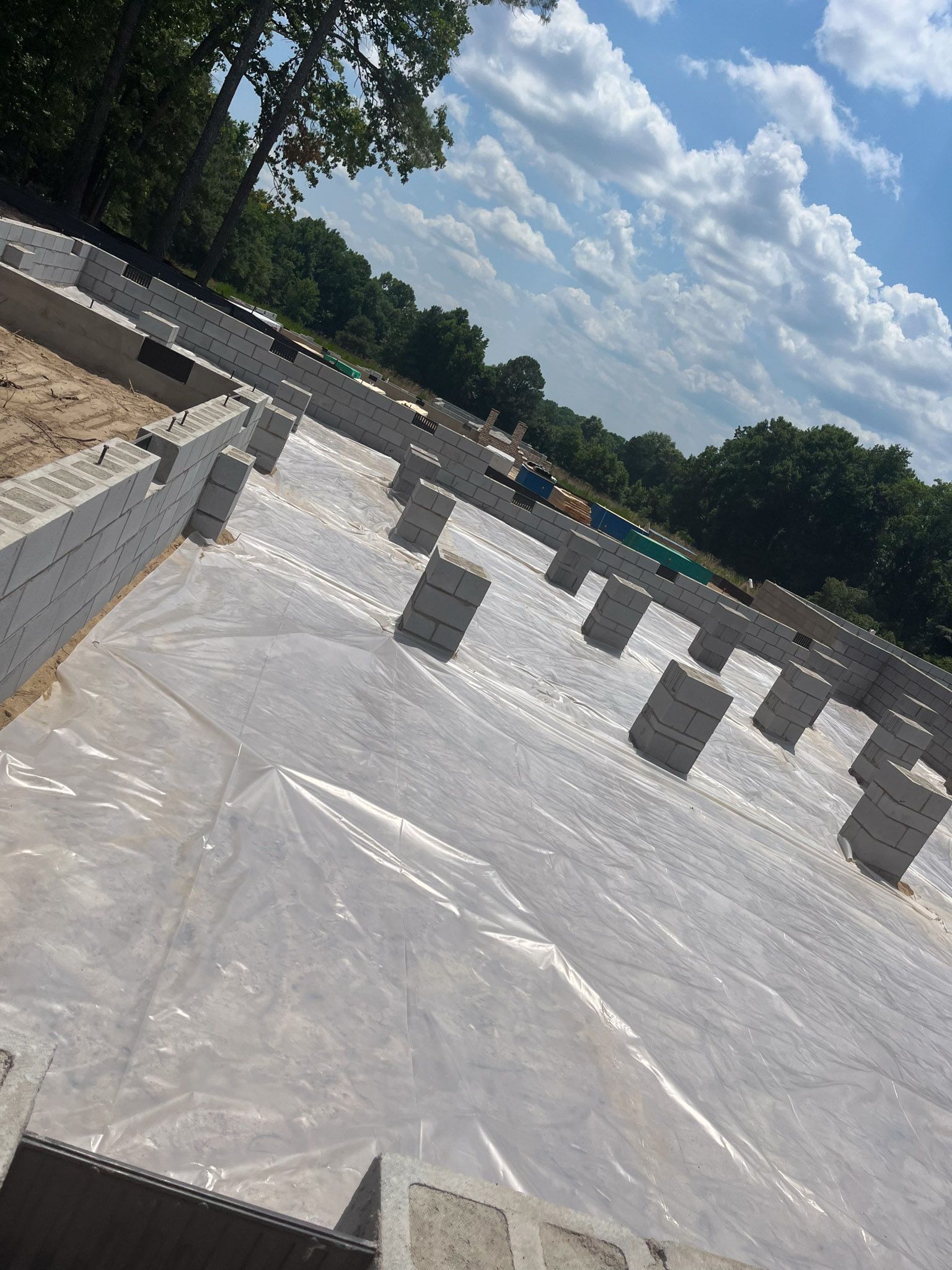 Construction site: foundation walls of white blocks, plastic sheeting, blue sky, trees in background.