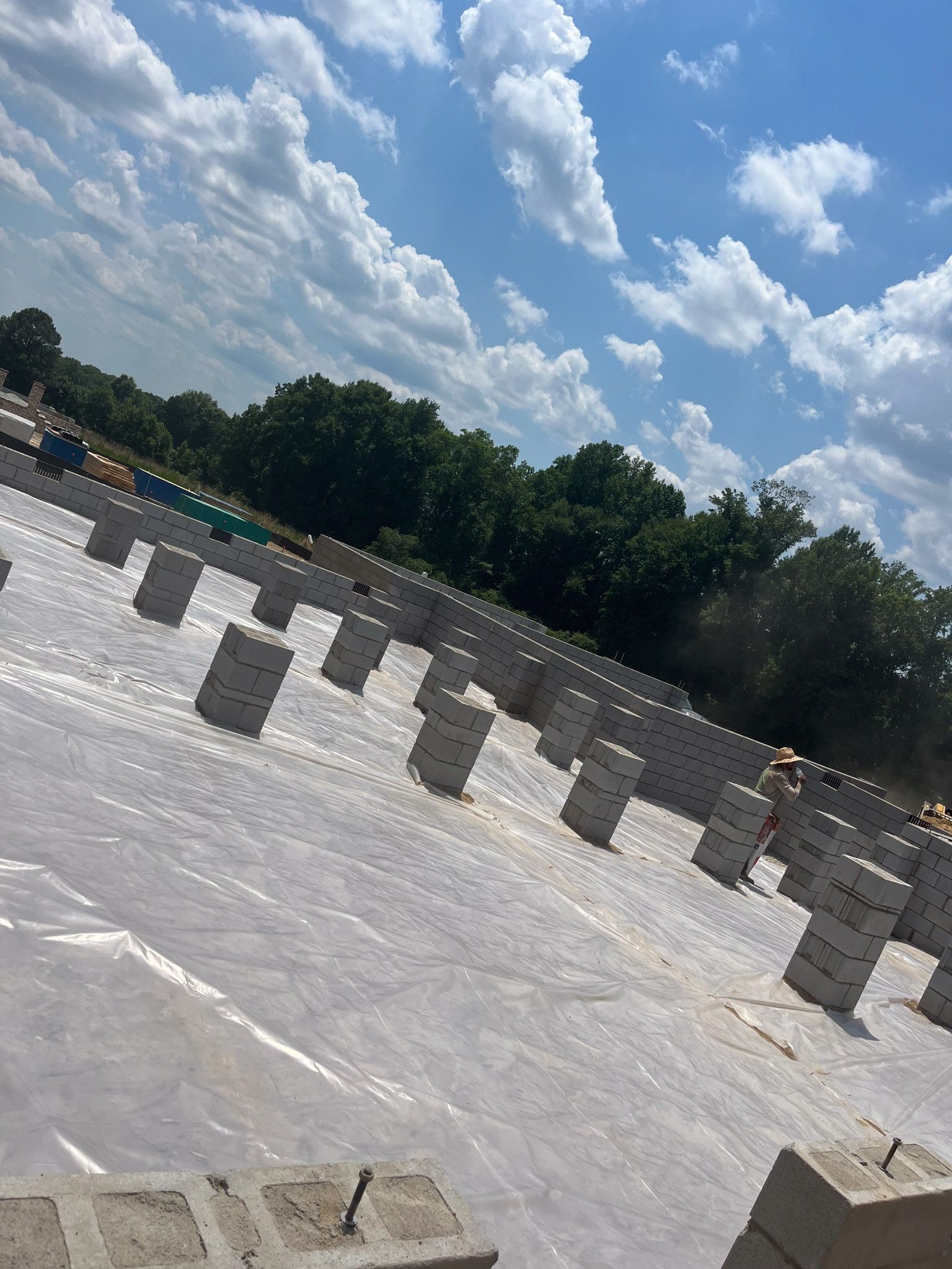Construction site with partially built brick structures on a covered surface; trees and blue sky background.