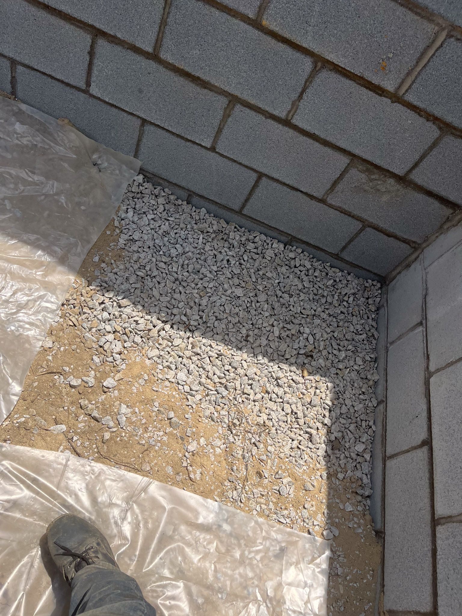 Gravel-filled space between gray brick wall and gray block foundation. Plastic sheeting visible, with a foot.